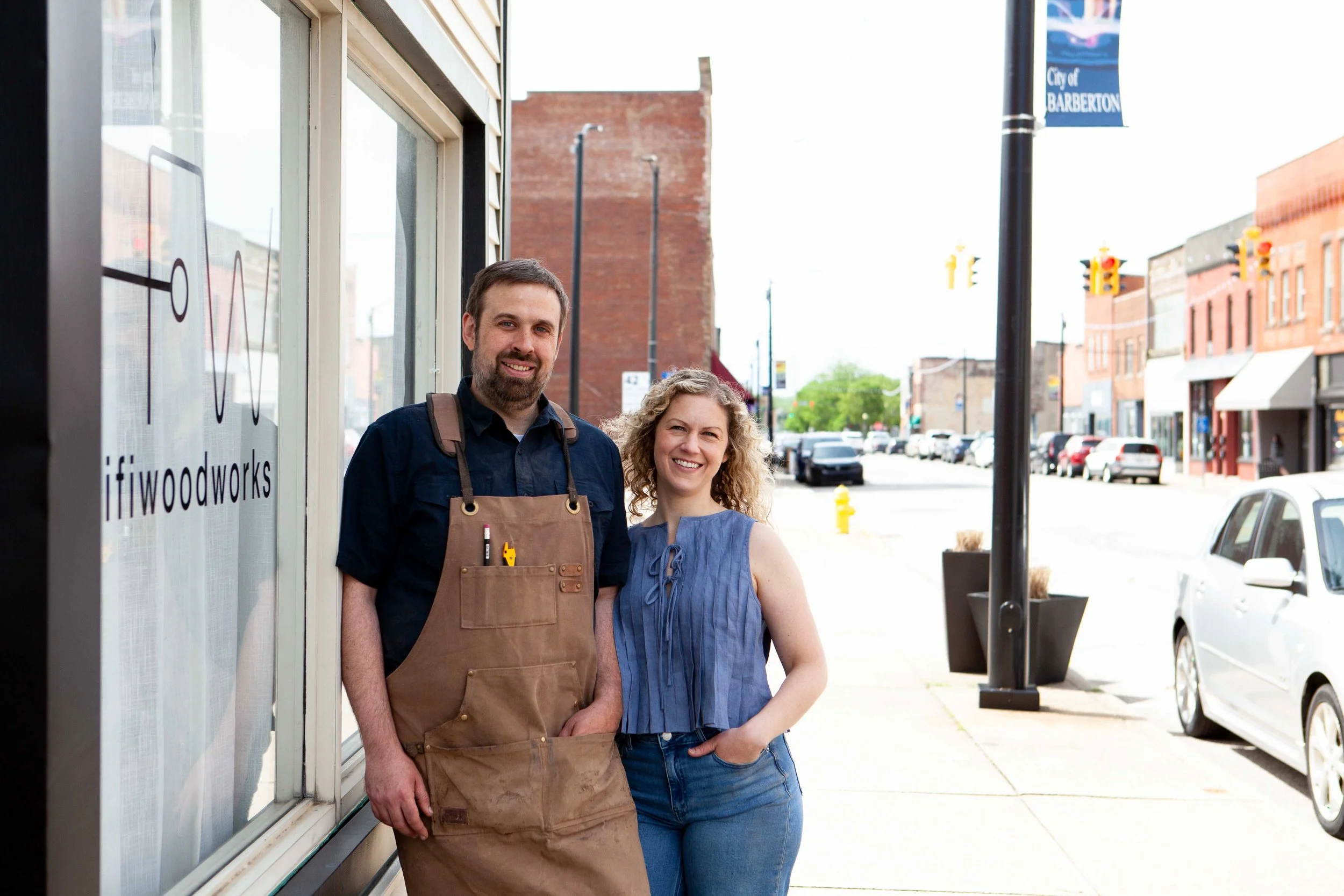 A man and woman standing outside a storefront on a city street, smiling at the camera. The man is wearing a dark shirt and an apron, the woman is wearing a sleeveless blue top. The storefront has a sign that reads 'Woodworks'.