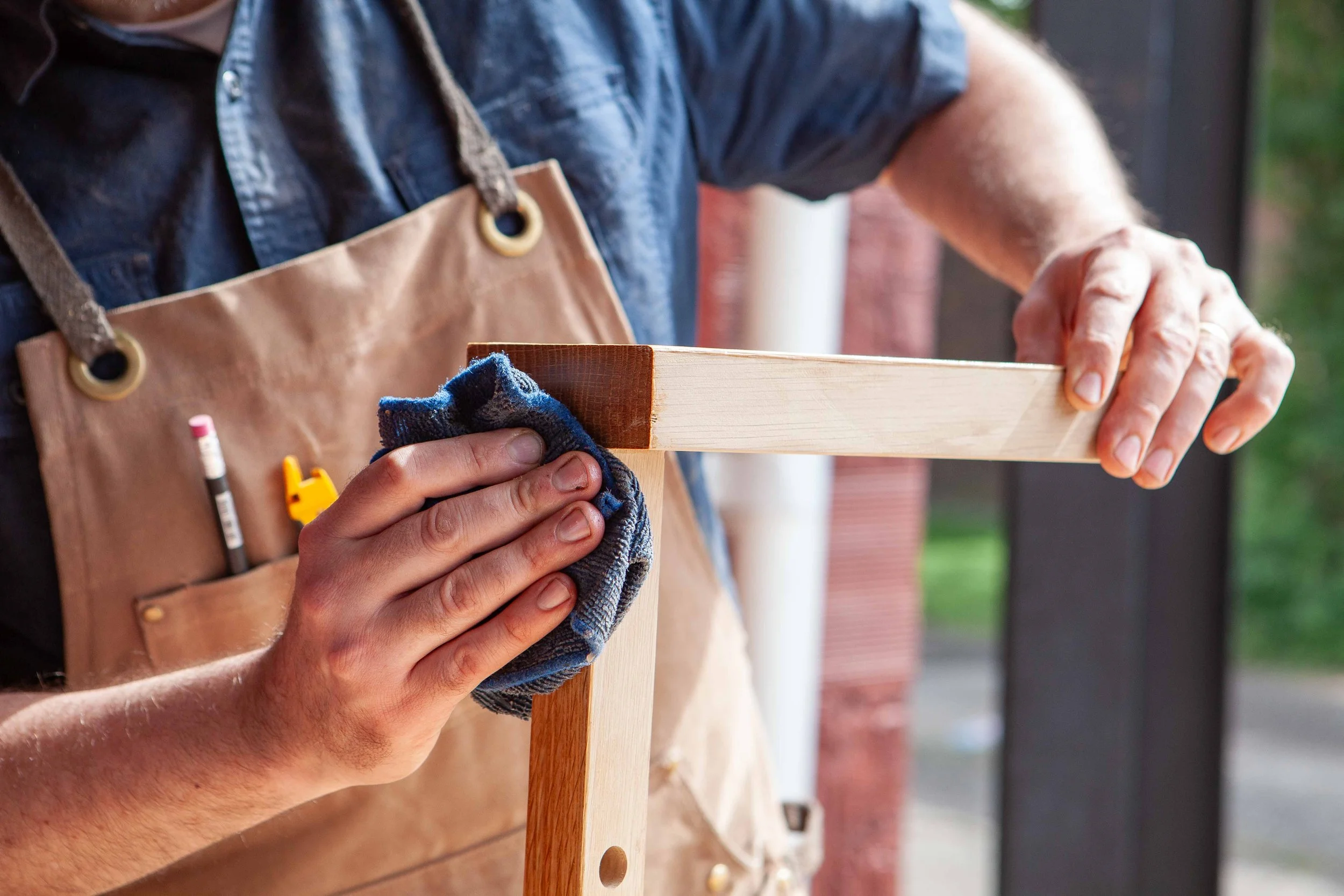 A person wearing a denim shirt and tan apron holding a wooden block and a cloth, working on a woodworking project outdoors.