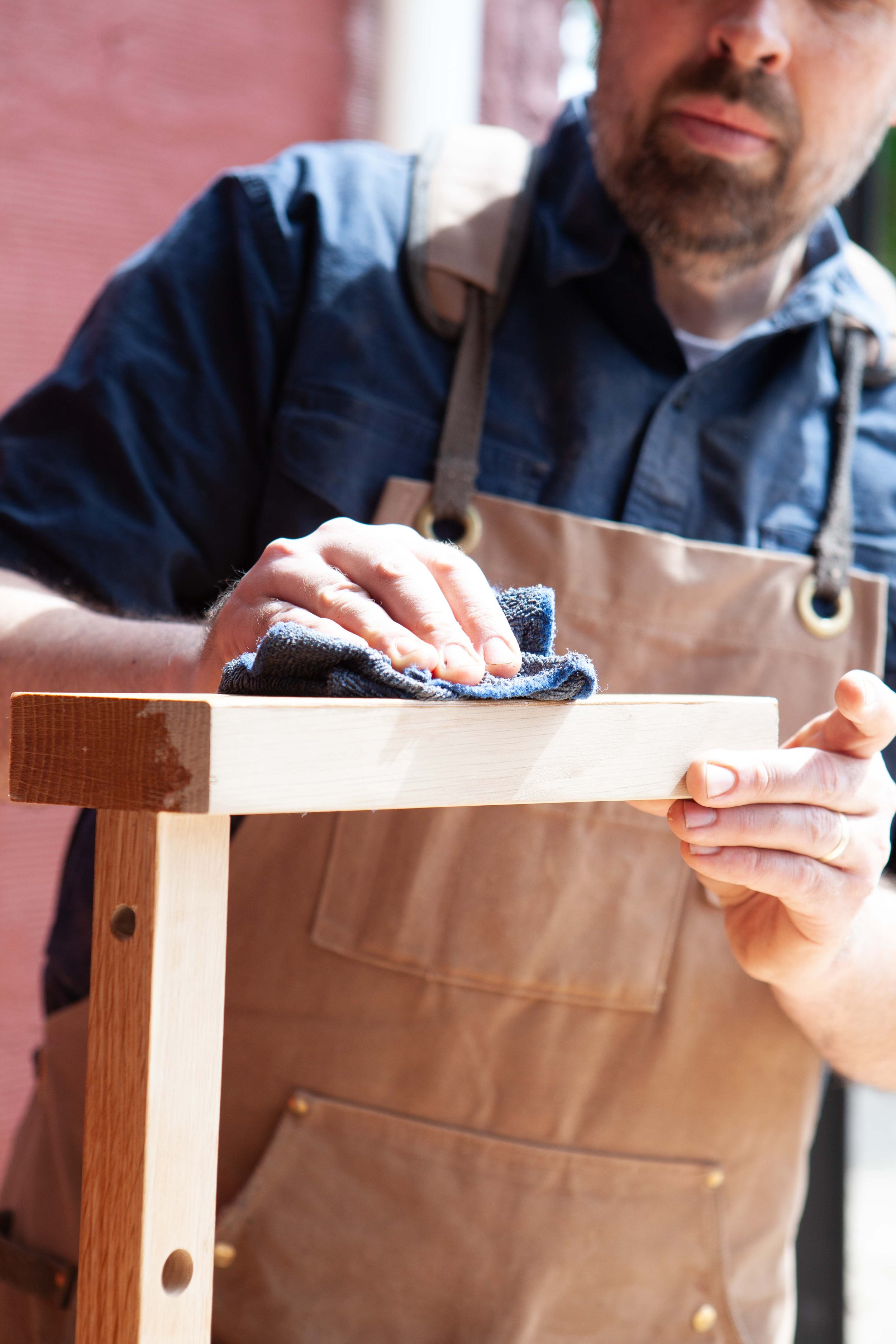 A person wearing a brown apron and a navy blue shirt is polishing a wooden surface with a cloth. The person's face is partially visible, showing a beard and mustache, and they are holding the cloth in their right hand. The focus is on the person's hand and the wooden surface, with a blurred background.