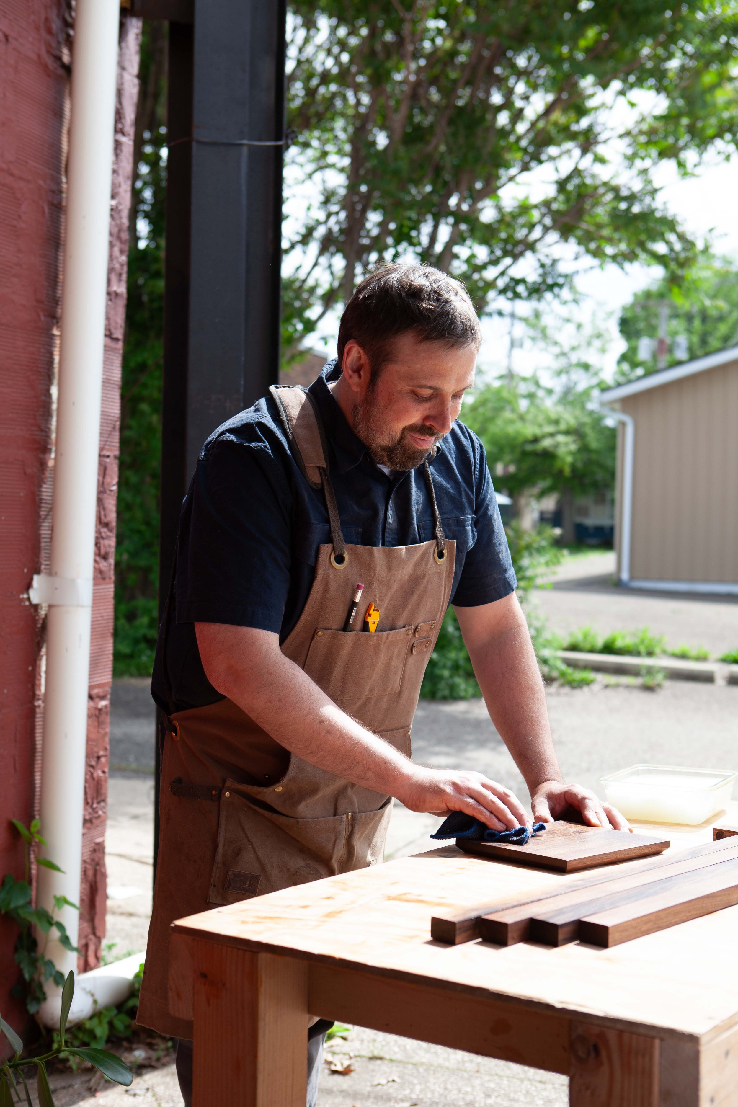 A man wearing a beige apron and dark shirt is working outdoors at a wooden table, sanding a piece of wood with a dark cloth. There are trees, a house, and other outdoor elements in the background.