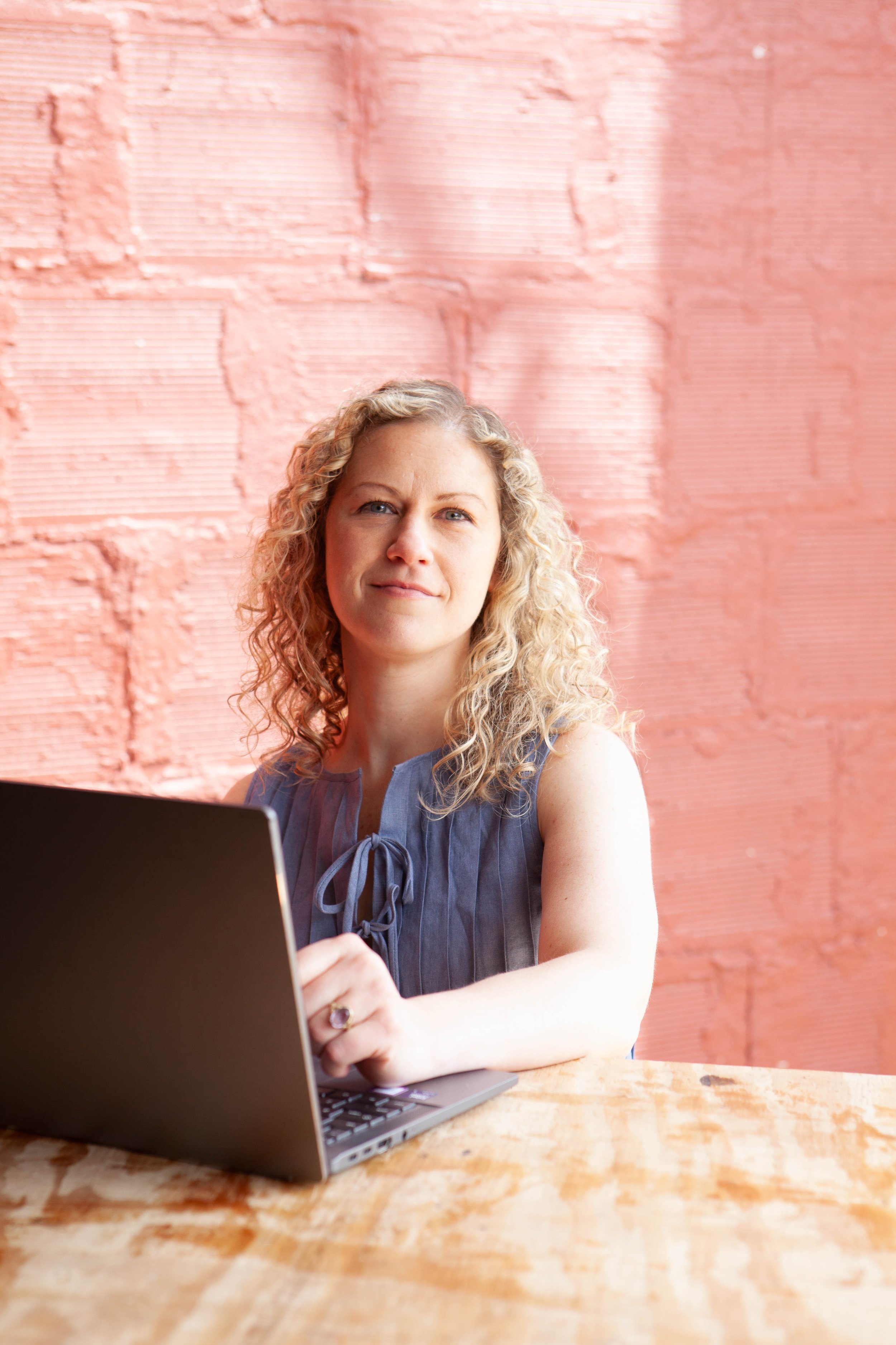 A woman with curly blond hair sitting at a wooden table with a laptop in front of her, against a pink brick wall background.