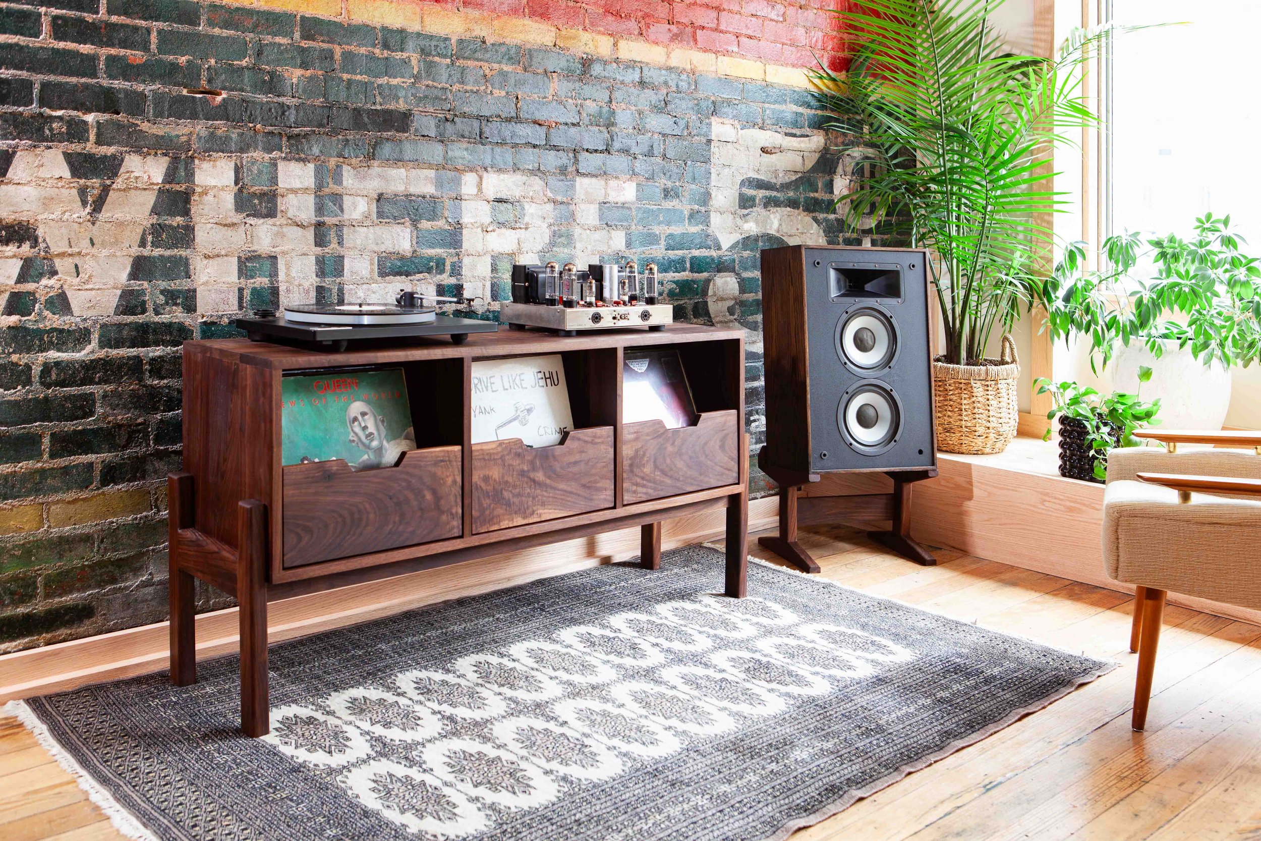Interior of a living room with a brick wall, a wooden sideboard holding a turntable, vacuum tube amplifier, and vinyl records, a large speaker, and plants near a window with natural light.