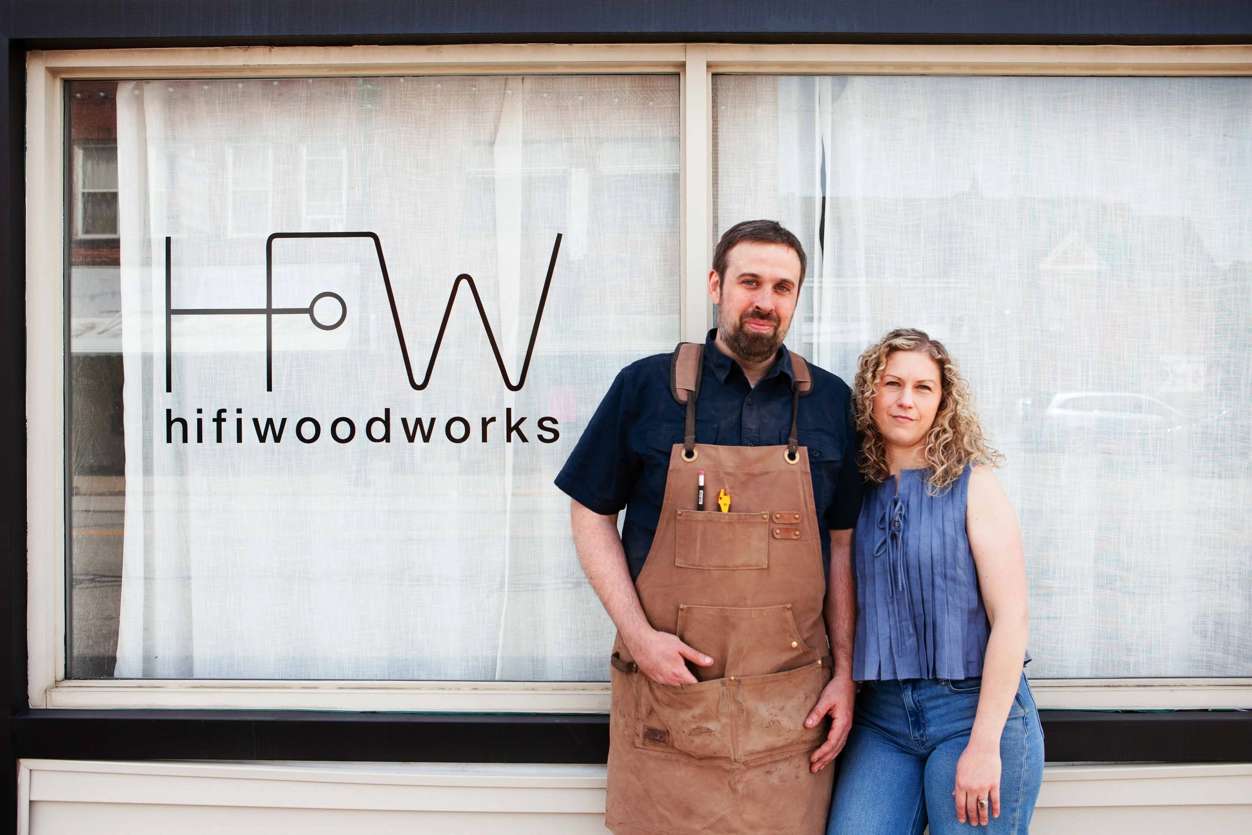 Two people, a man with a beard wearing a dark shirt and an apron, and a woman with curly blonde hair wearing a sleeveless top, standing in front of a window with the words 'hifiwoodworks' and an abstract logo.