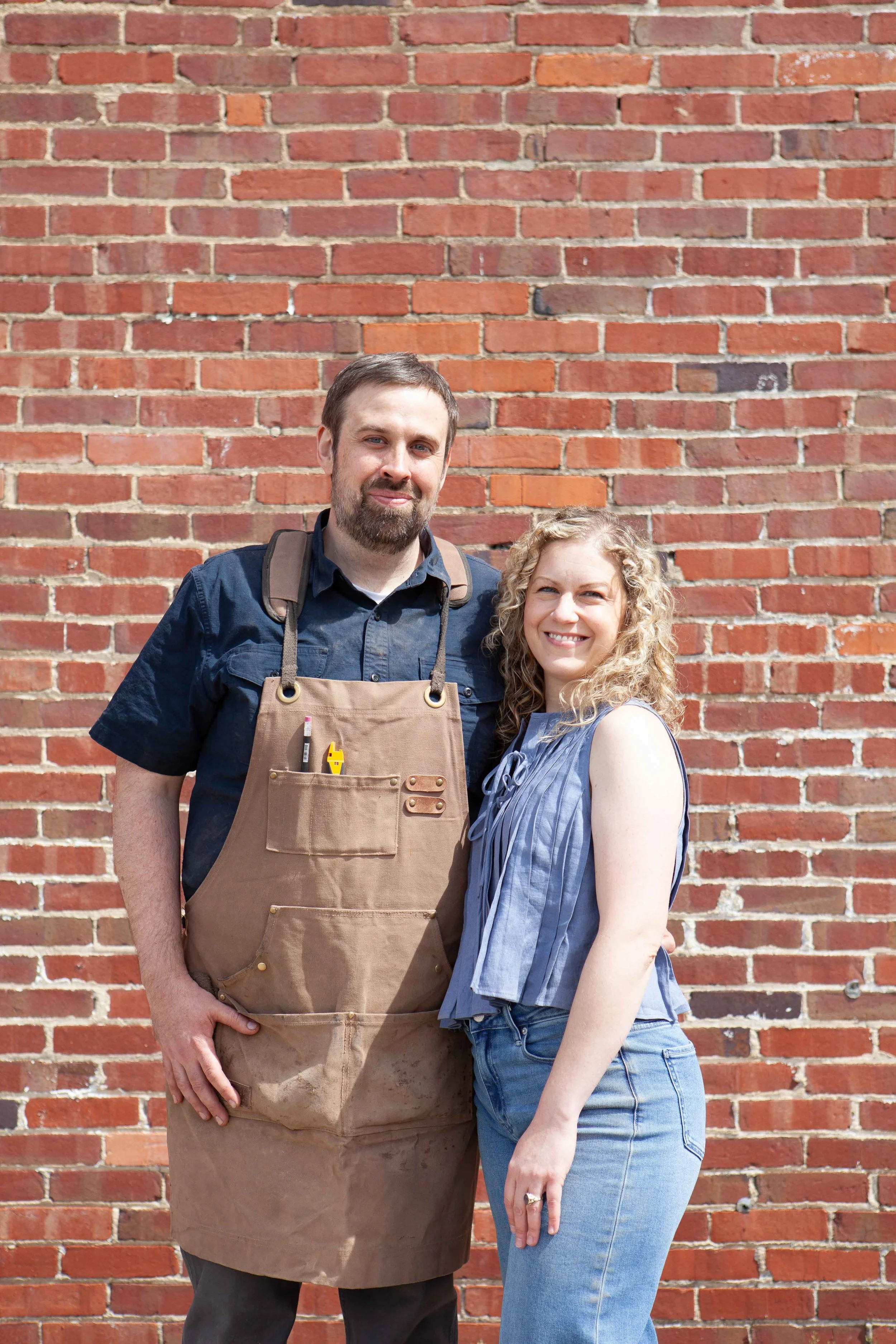 A man with brown hair and beard wearing a dark blue shirt and brown apron standing next to a woman with curly blonde hair wearing a sleeveless top and jeans, both smiling, in front of a red brick wall.