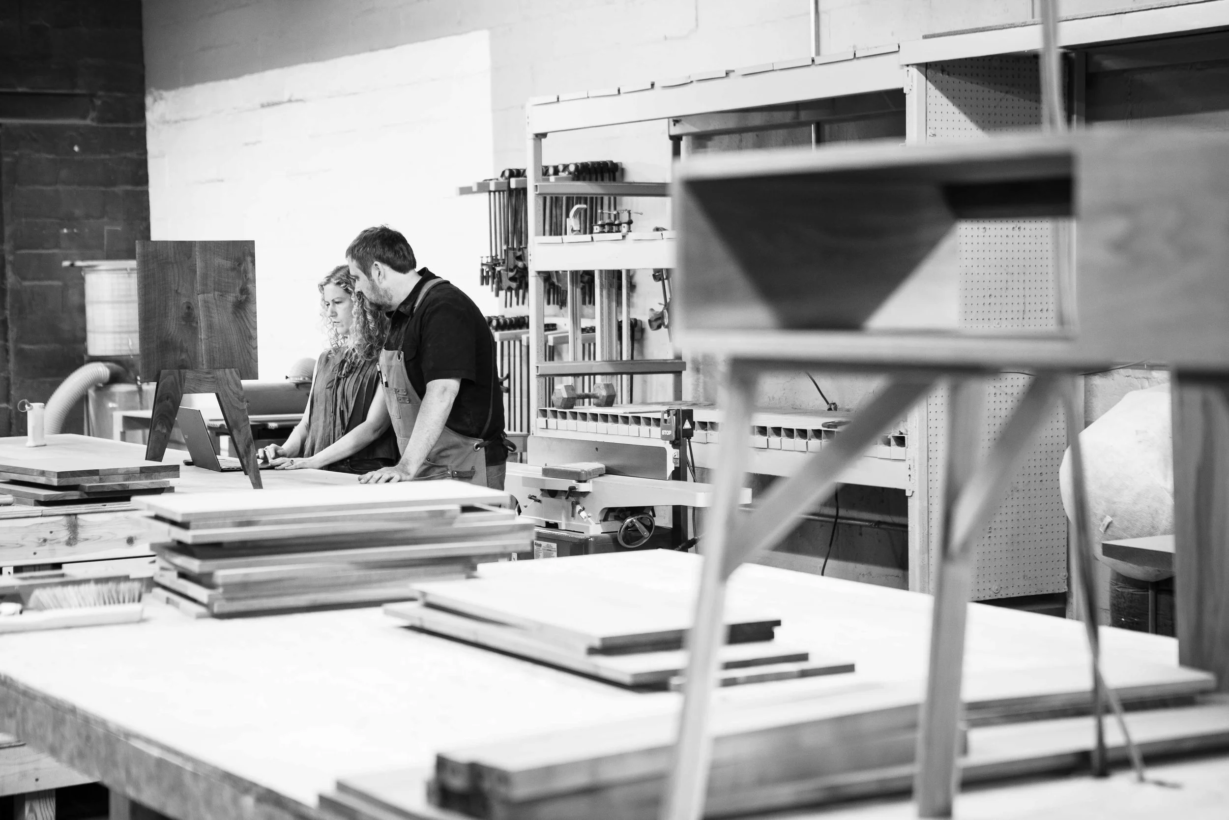 Two people working together in a woodworking shop, examining a computer monitor among sawhorses and stacks of wooden boards.