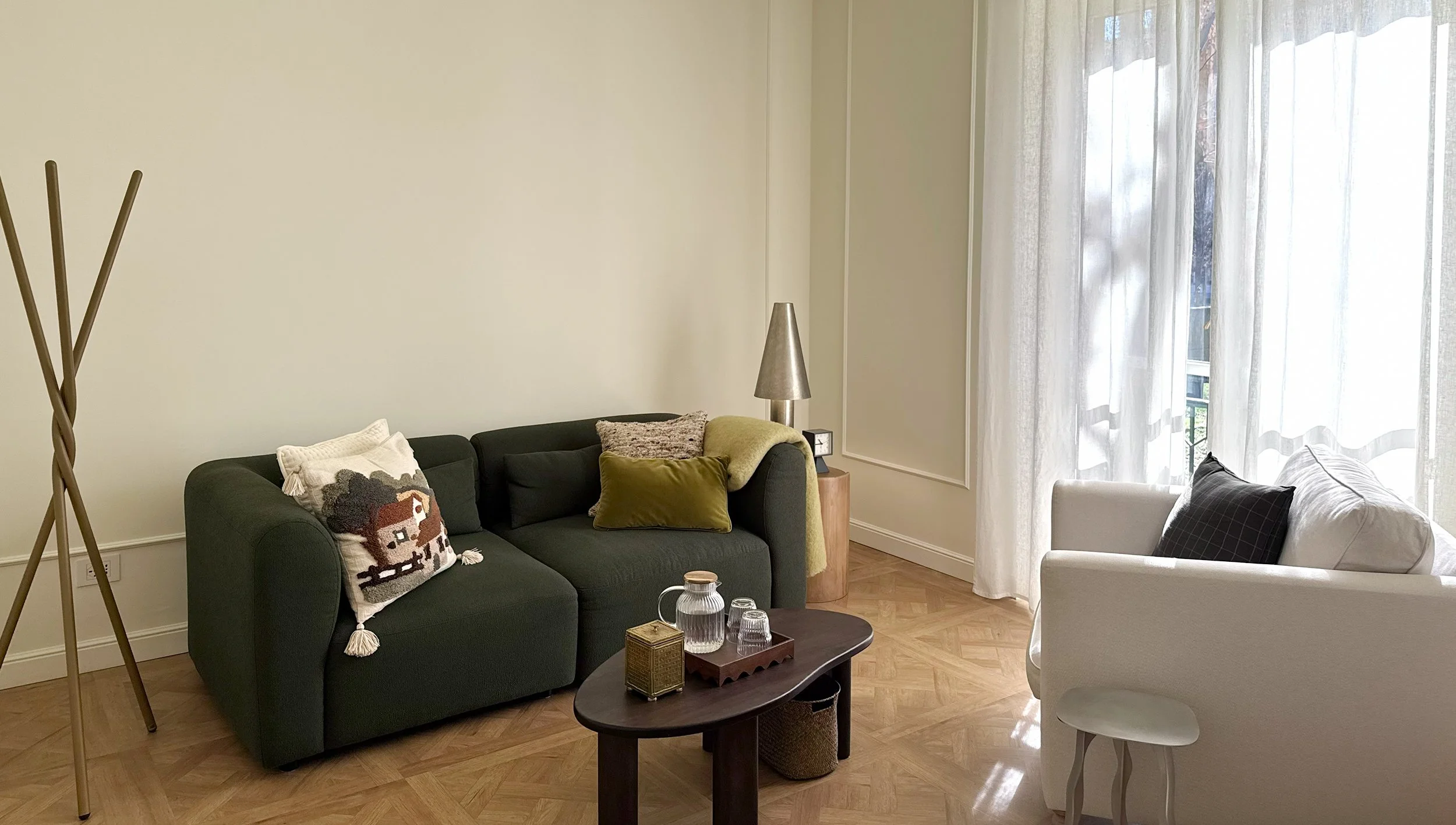 Studio psicologico con divano nero decorato con cuscini, tavolino rotondo in legno con bicchieri e oggetti decorativi, lampada da tavolo, divano bianco, grandi finestre con tende trasparenti, pareti chiare e pavimento in parquet