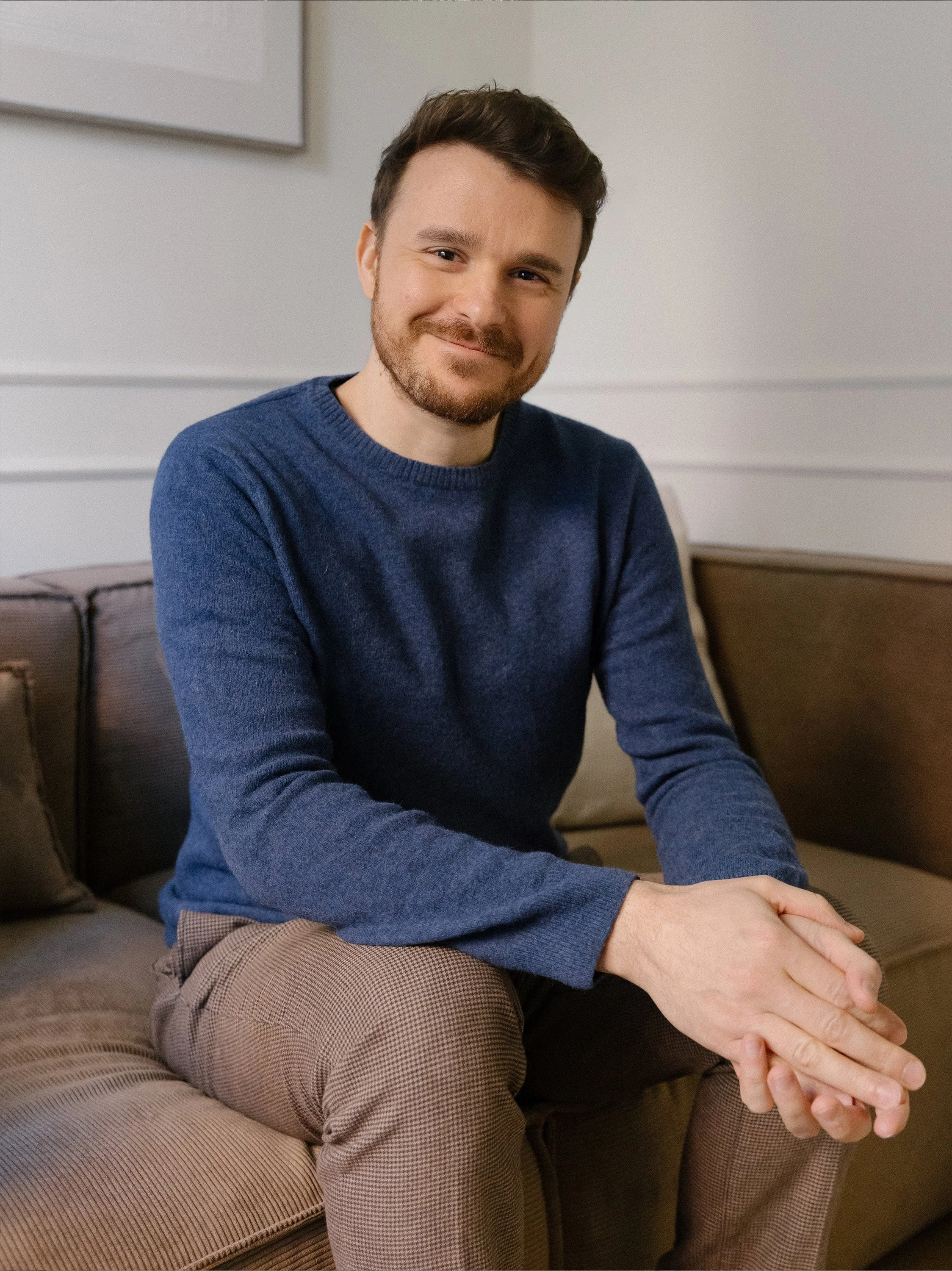 Psicologo seduto su un divano, sorridente, con indumenti casual beige e blu, capelli corti e barba, in un ambiente accogliente.