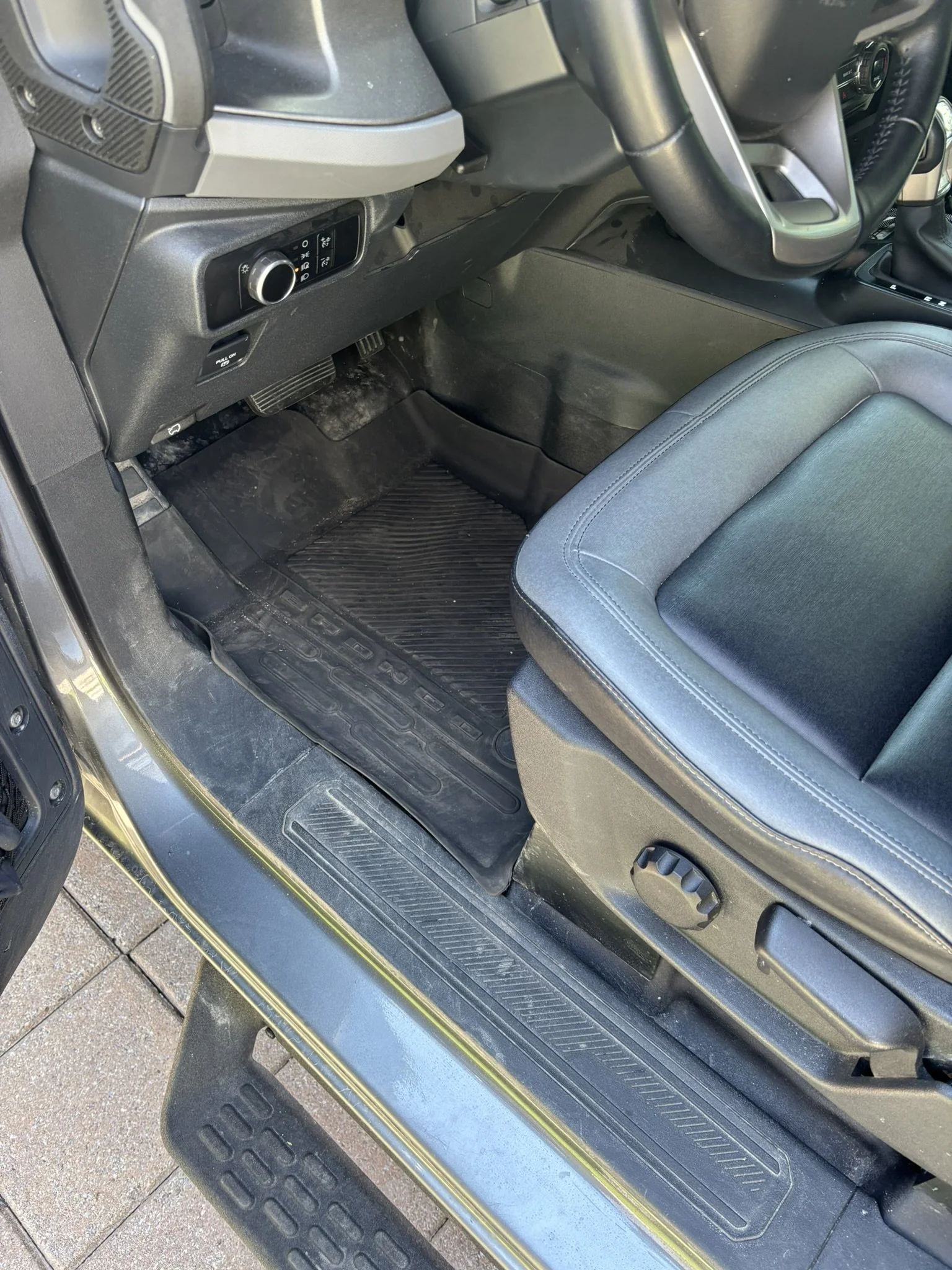 The interior of a vehicle shows the driver's side floor area, including the foot pedals, floor mat, and part of the seat. The dashboard features various controls and a rotary switch; the steering column and part of the gear shifter are also visible.