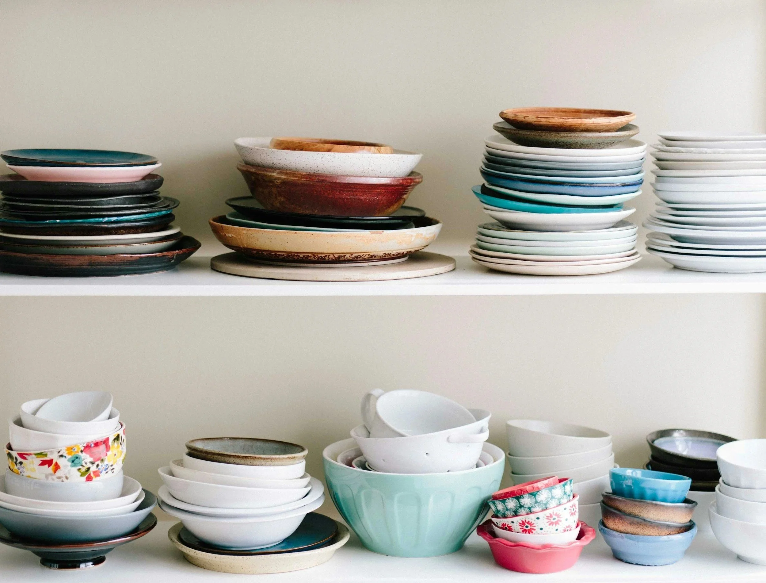 Stacks of various colorful ceramic bowls and plates on a white shelf against a white wall.