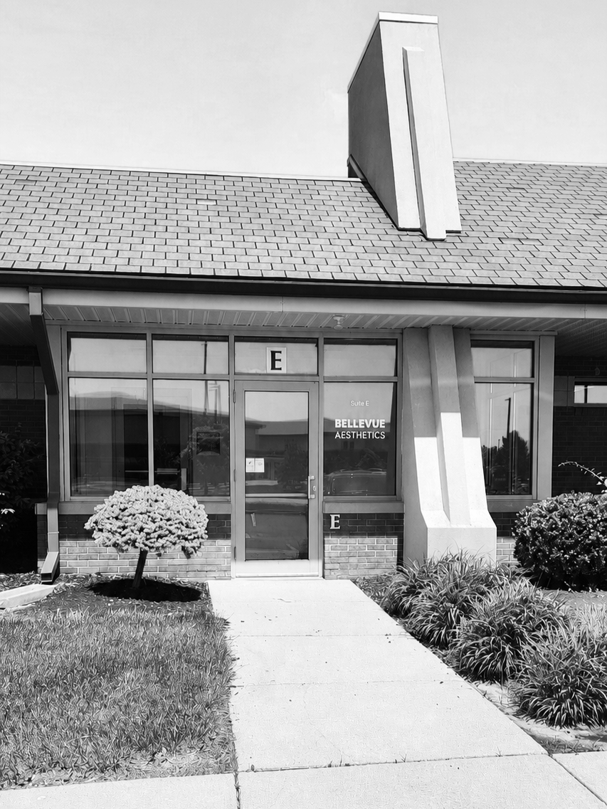The exterior of a building with large glass windows and a door, featuring a sign that reads 'Bellevue Aesthetics.' There is a small landscaped area with shrubs and a concrete sidewalk leading to the entrance.