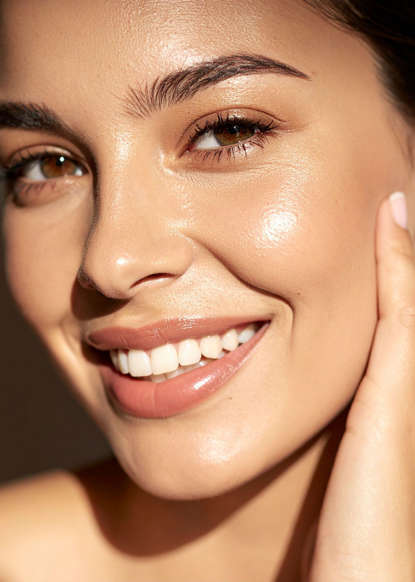 Close-up of a smiling woman with brown eyes, full lips, clear skin, and dark brown hair. She has a natural makeup look with subtle eye makeup and light lipstick.