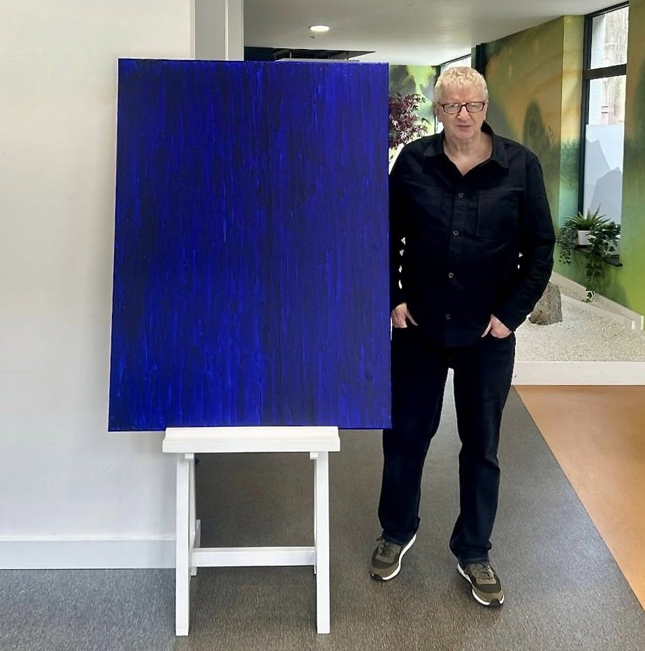Contemporary Irish artist Paul Bell standing next to a large, dark blue abstract painting, THE BLUE, on an easel indoors.