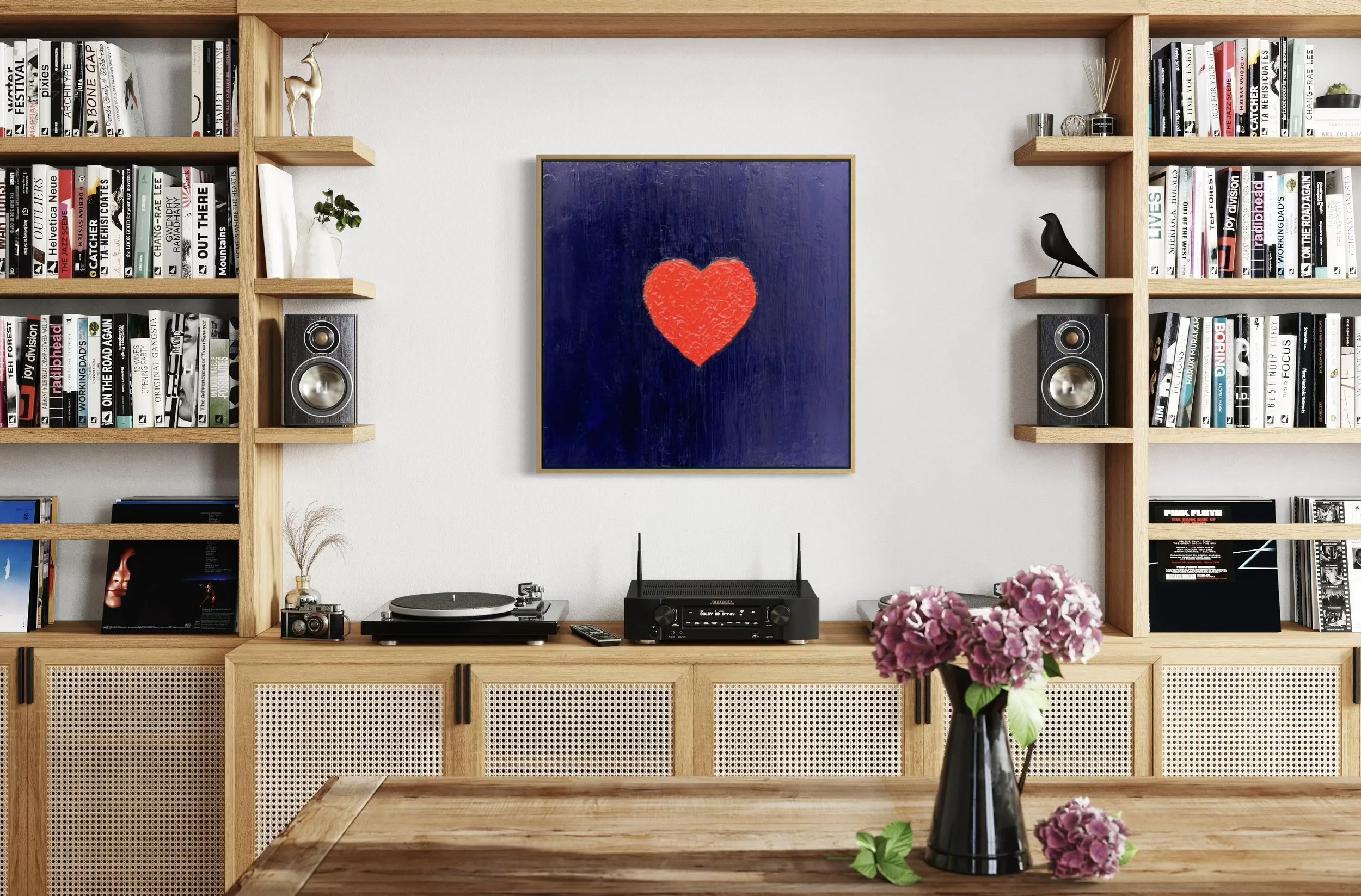 Living room wall with built-in wooden bookshelves, a framed artwork, FOR YOU by Paul Bell, a red heart on a purple background, and various decorations. A table with a flower vase and pink hydrangeas.