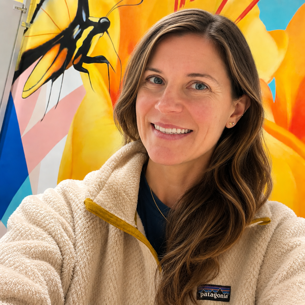 A woman with long, wavy brown hair and blue eyes smiling for a selfie in front of a colorful mural featuring a bee, orange, yellow, pink, blue, and black abstract shapes.