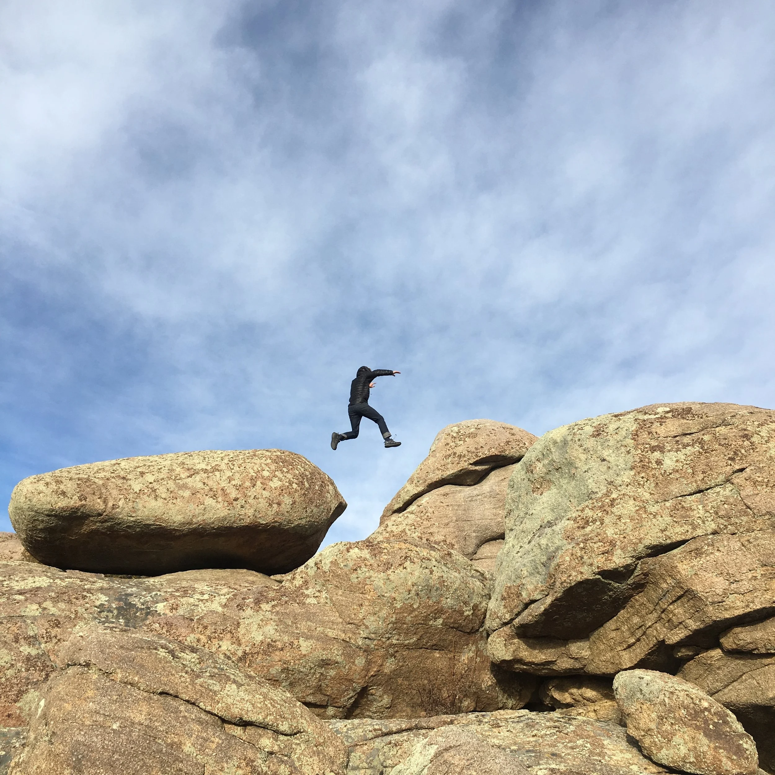 A person is mid-jump between rocks in a natural outdoor setting, with a cloudy sky in the background.