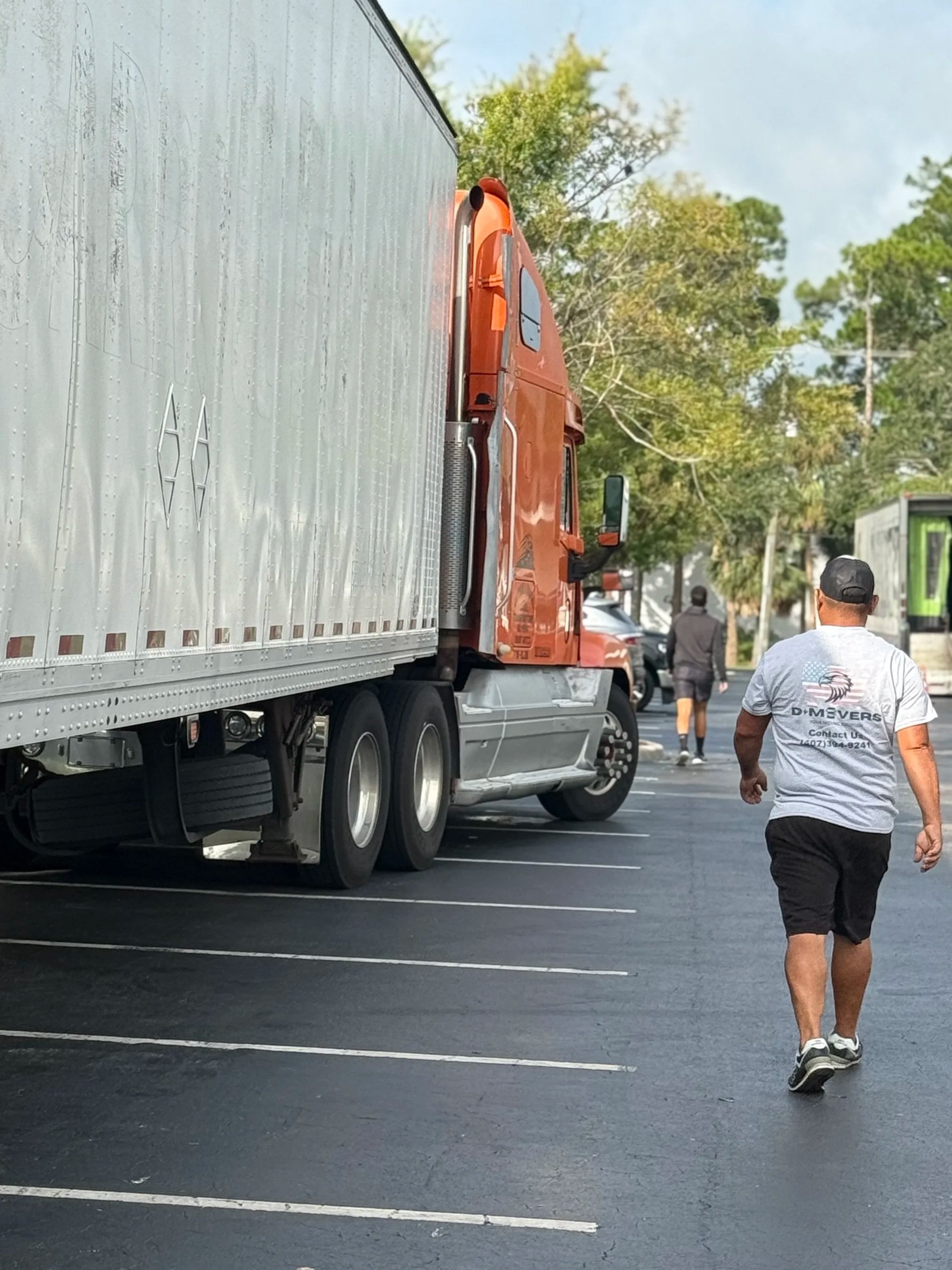 A man walking in a parking lot near a large orange semi-truck with a silver trailer. The man is wearing a white t-shirt with a logo on the back, black shorts, and sneakers. The background shows trees and a street with some cars.
