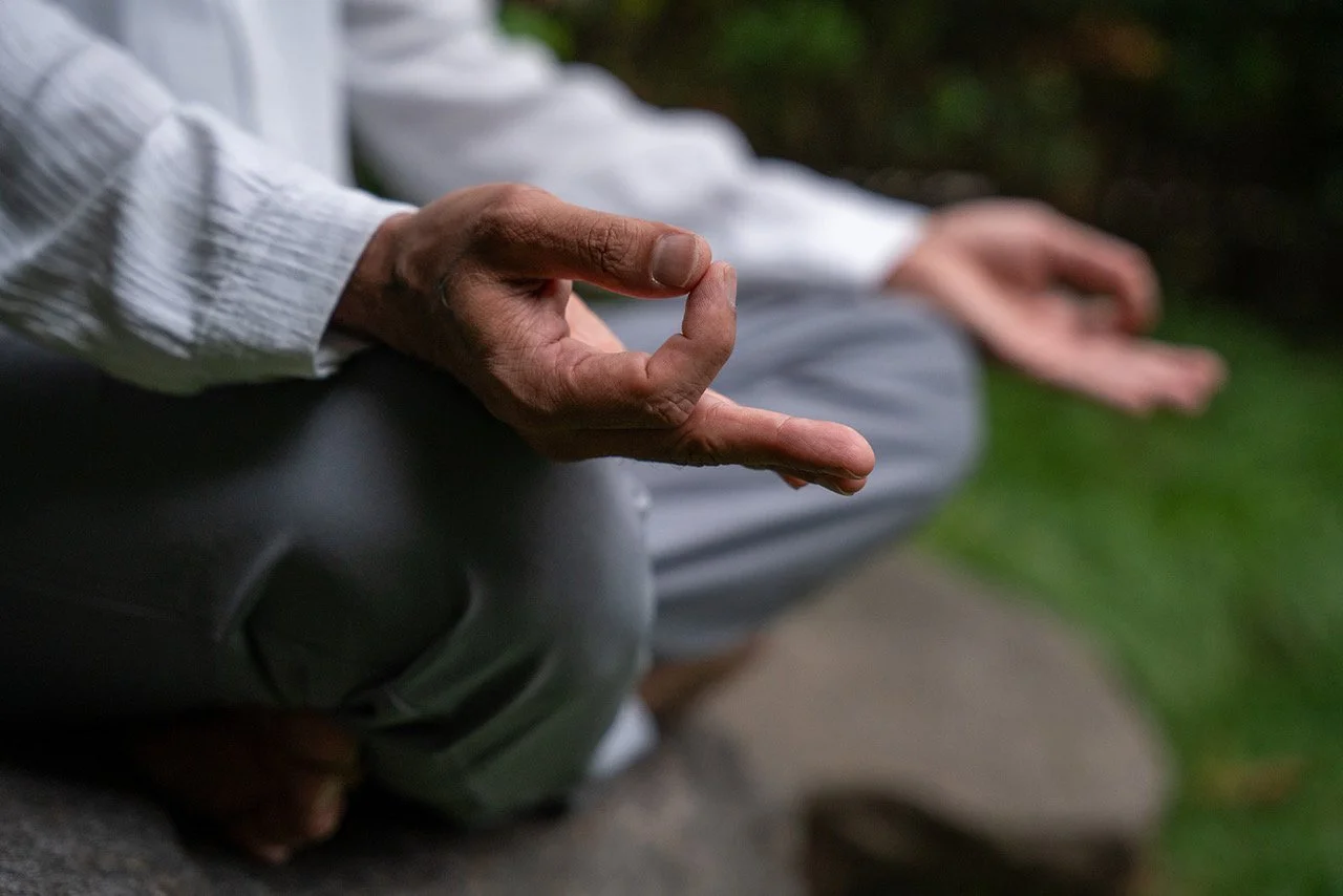 Close-up of hands in a meditation pose, with the thumb and index finger touching, sitting outdoors on a stone surface with greenery in the background.