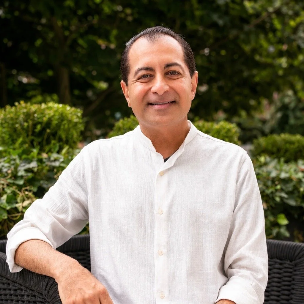 A man smiling outdoors in front of green bushes, wearing a white button-up shirt with the sleeves slightly rolled up.