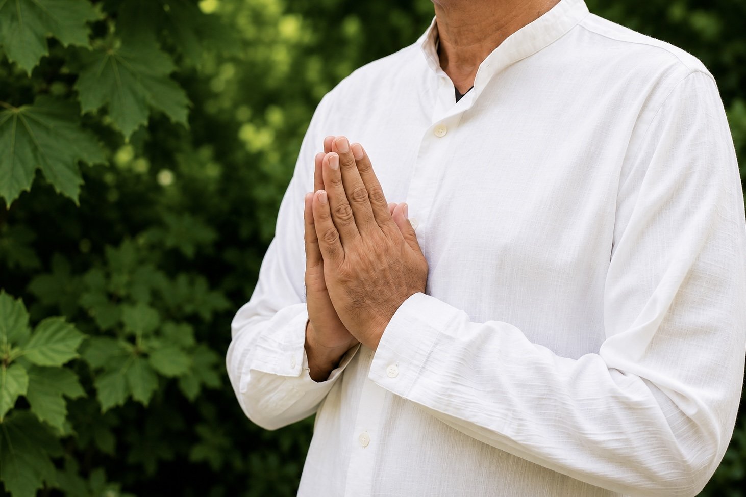 A man in a white shirt standing outdoors with green foliage in the background, hands pressed together in a prayer-like gesture.