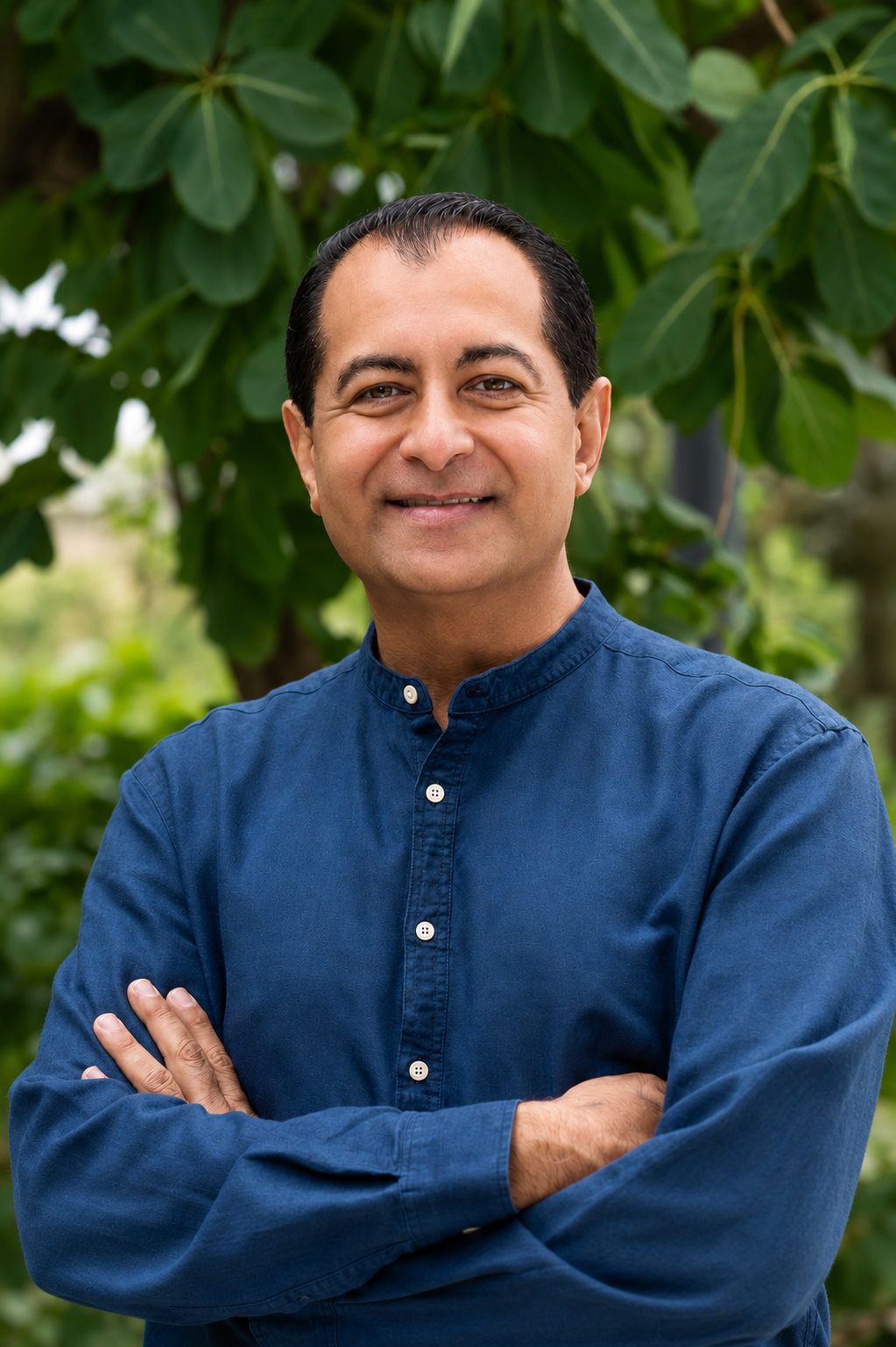 A man with short dark hair, wearing a navy blue button-down shirt, standing outdoors in front of large green leaves, smiling with arms crossed.