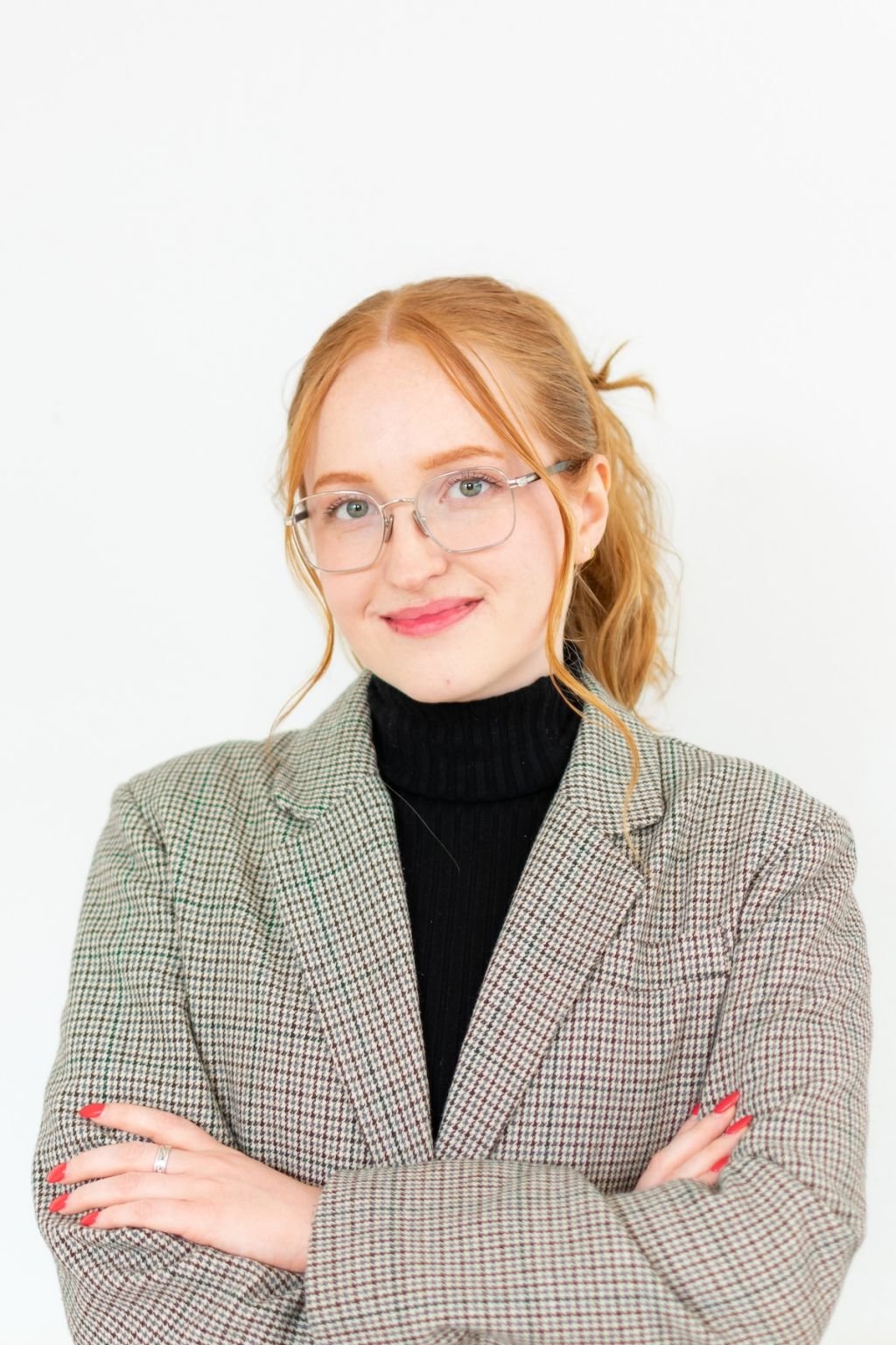 Portrait of a young woman with red hair, glasses, wearing a black turtleneck and a checked blazer, standing against a white background.