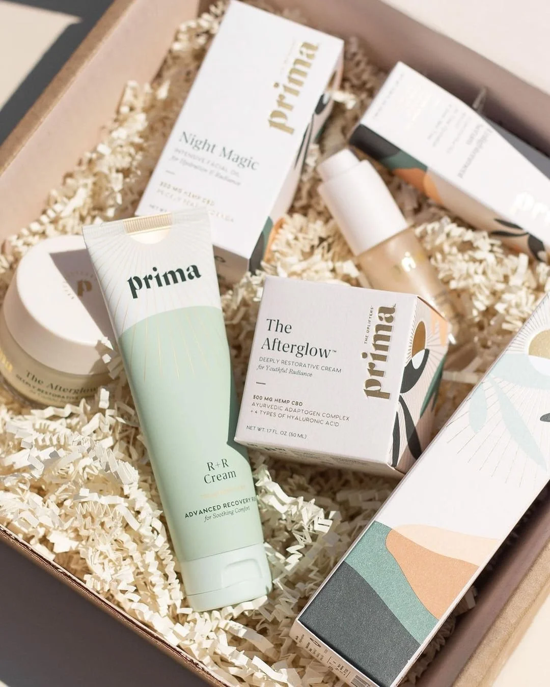 A gift box filled with skincare products, including Prima skincare items like R+R Cream and The Afterglow, cushioned with shredded paper for packaging.