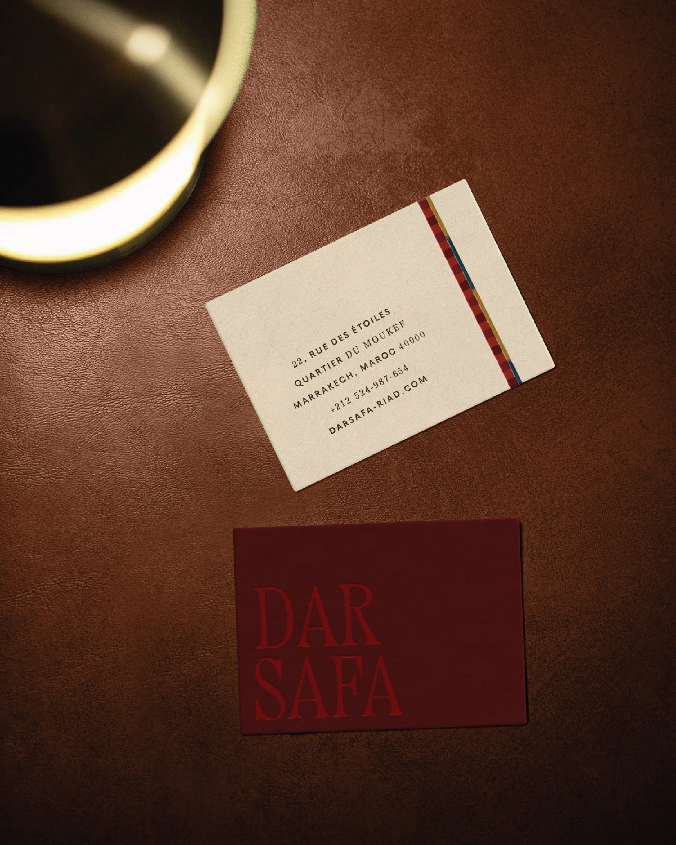 A business card with an address in Morocco, a phone number, and a website, placed on a brown textured surface, with part of a dark red book with red text visible at the bottom, and a cup of coffee in the top left corner.
