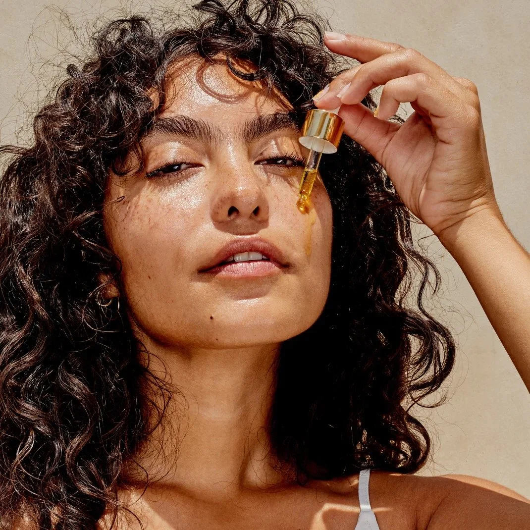 A woman with curly hair using a dropper to apply facial oil to her face.