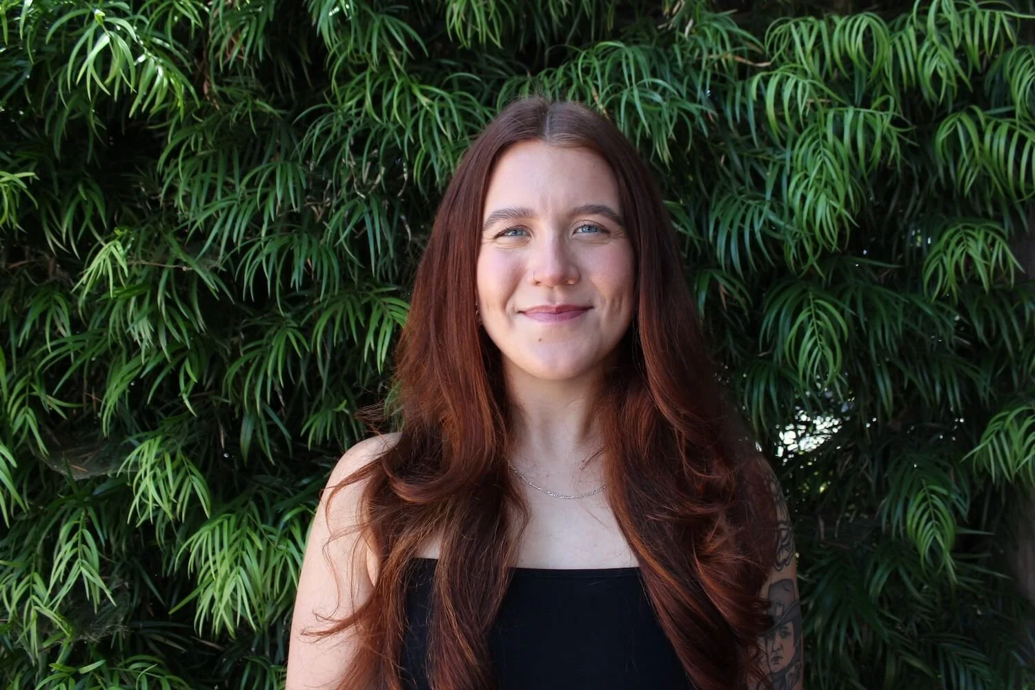 A smiling woman with long, wavy, reddish-brown hair standing in front of dense green foliage.