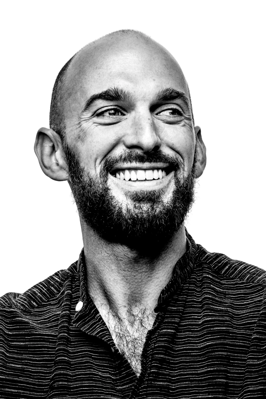 A smiling man with a beard and bald head, wearing a striped shirt, looking to his right against a plain white background.