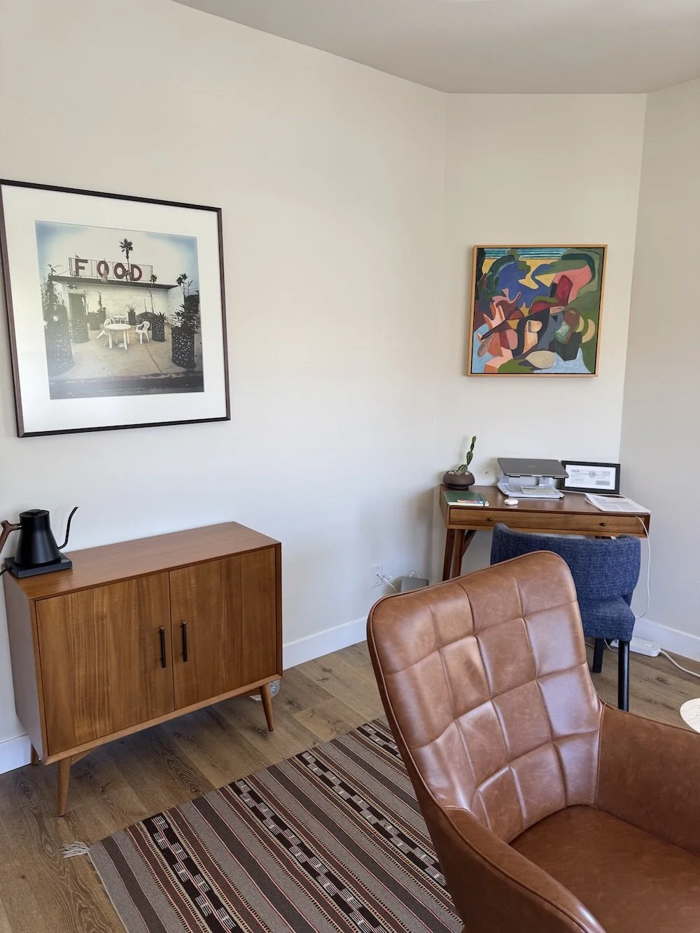 Corner of a room with a wooden cabinet, a leather armchair, a desk with a printer, a framed colorful abstract painting, and a black and white framed photo of an outdoor food stand with chairs and a table.