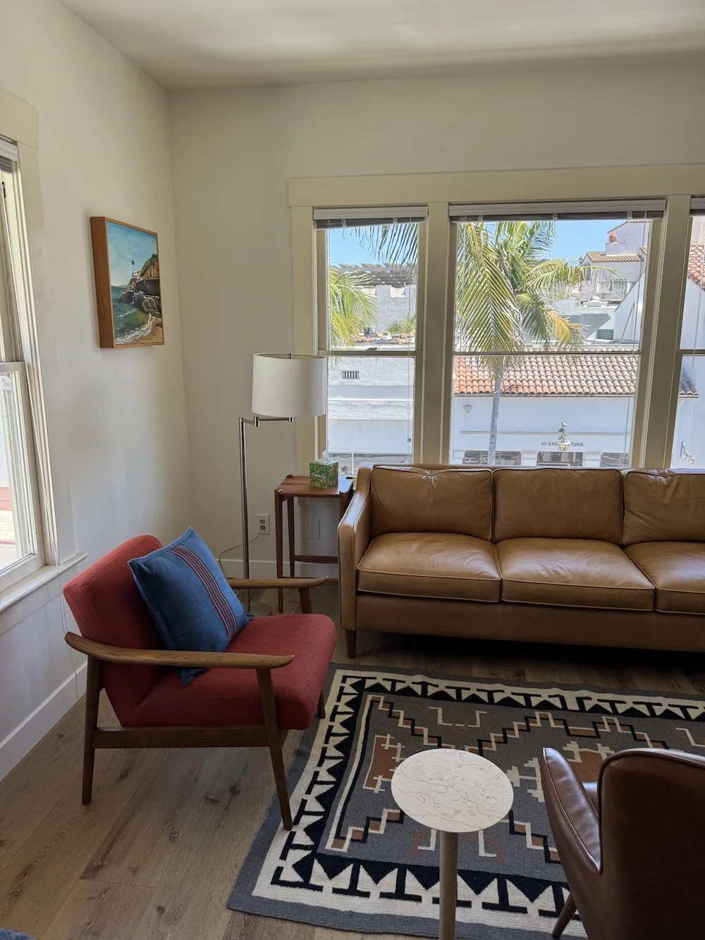 Living room with a tan leather sofa, a red armchair with a blue pillow, a wooden side table with a lamp and a tissue box, a small round table, a patterned rug, and two large windows showing palm trees and rooftops outside.