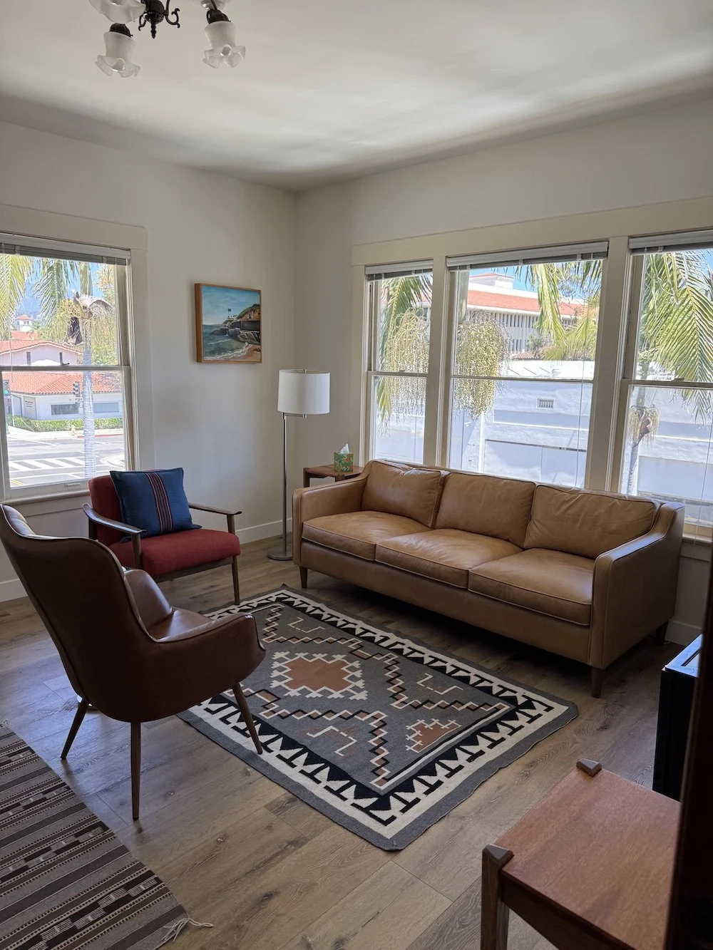 Bright living room with large windows, tan sofa, red armchair, brown leather chair, and colorful geometric rug.
