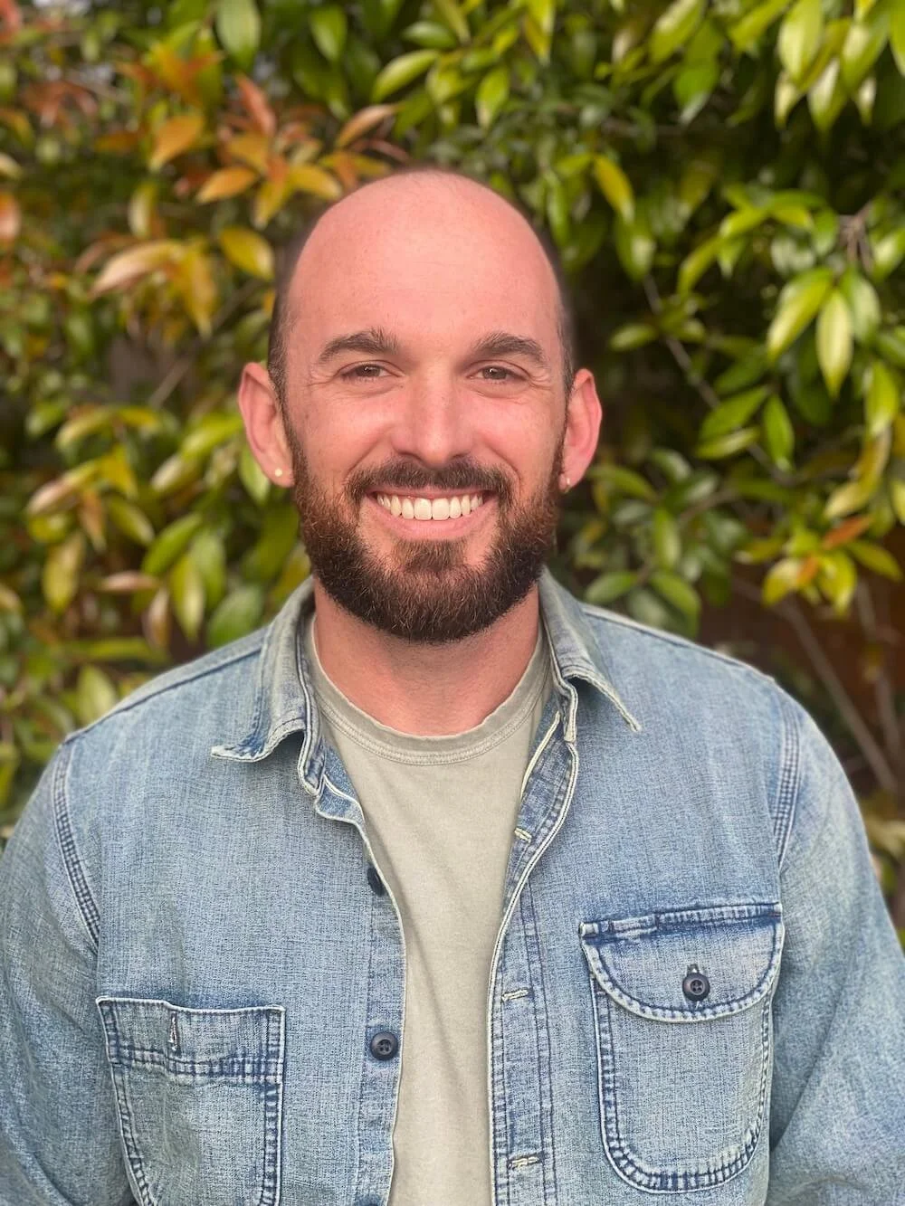 A smiling man with a beard and no hair wearing a denim jacket and a light-colored shirt standing in front of green leafy bushes.
