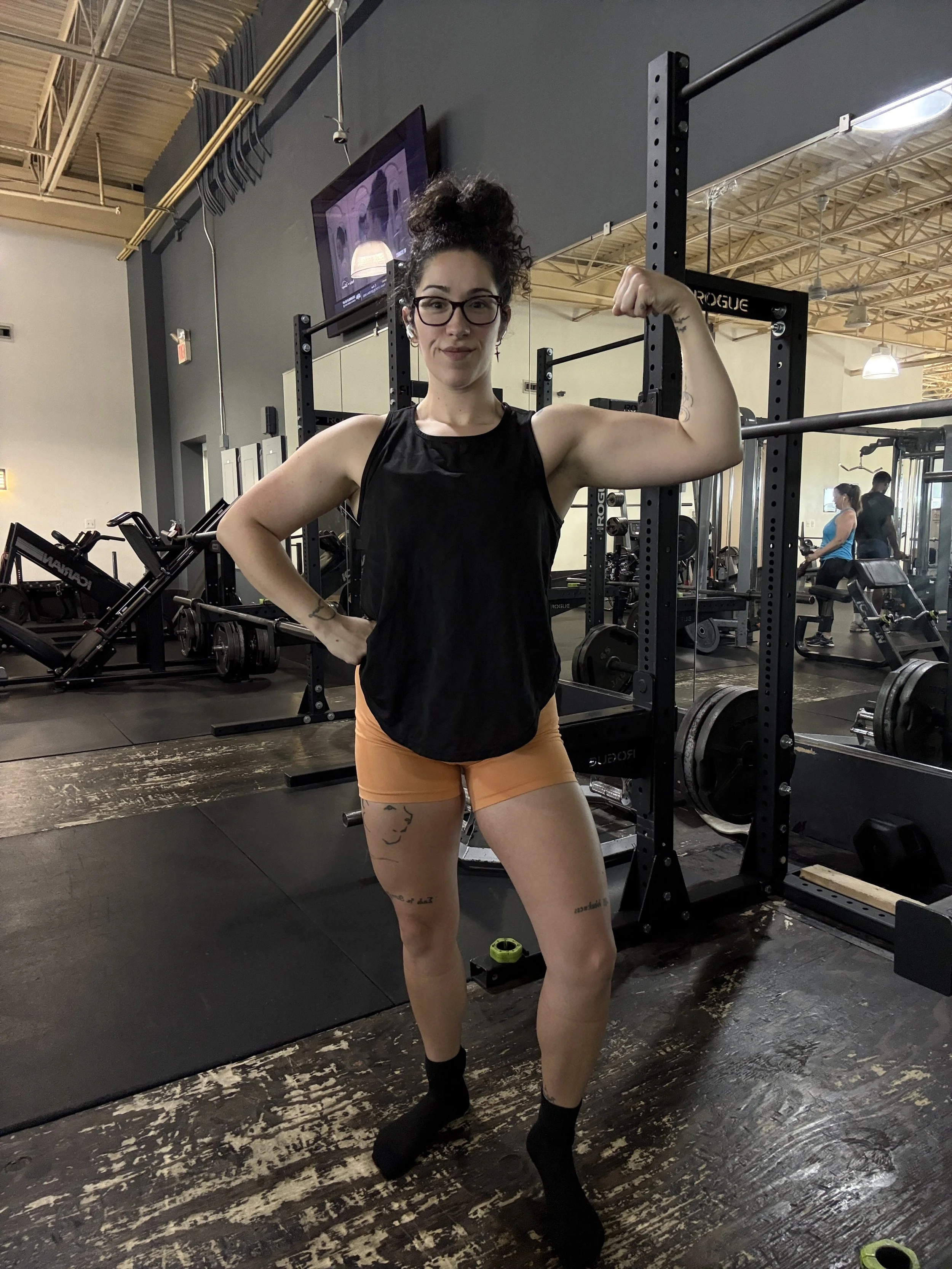 A woman with glasses and curly hair in a top and shorts flexing her right arm in a gym.
