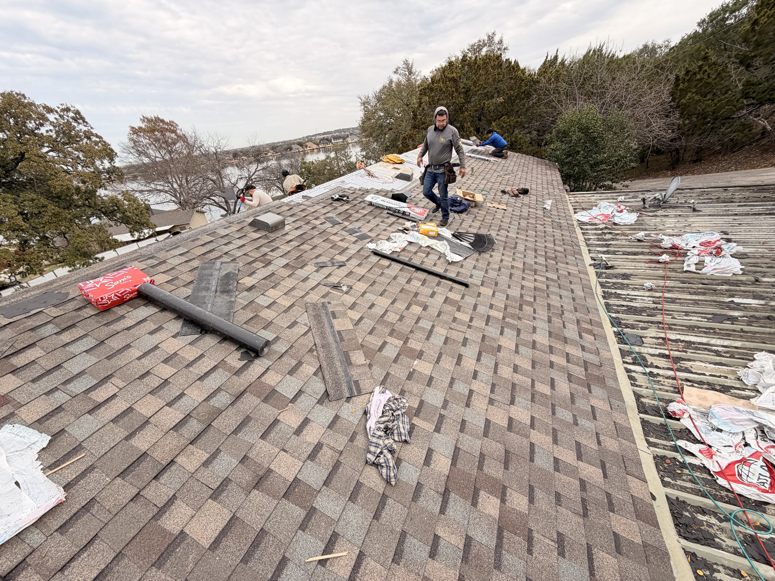 People working on a roof with shingles, tools, and materials scattered around, some sitting and others standing, with trees and a view of a lake or river in the background. granbury tx