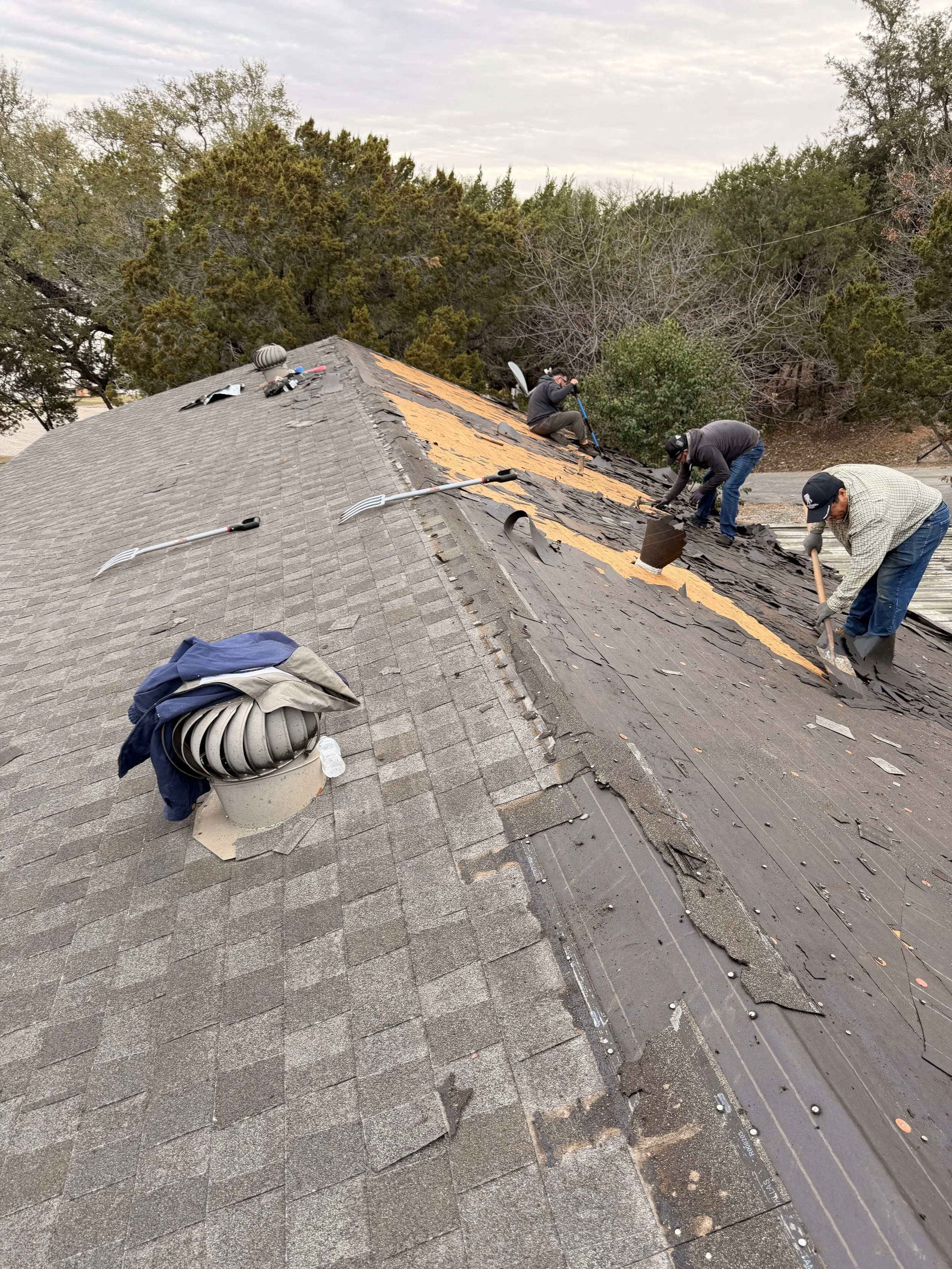 Workers repairing a damaged section of a house roof with gray shingles, using tools and scattered roofing materials, amidst trees and cloudy sky.