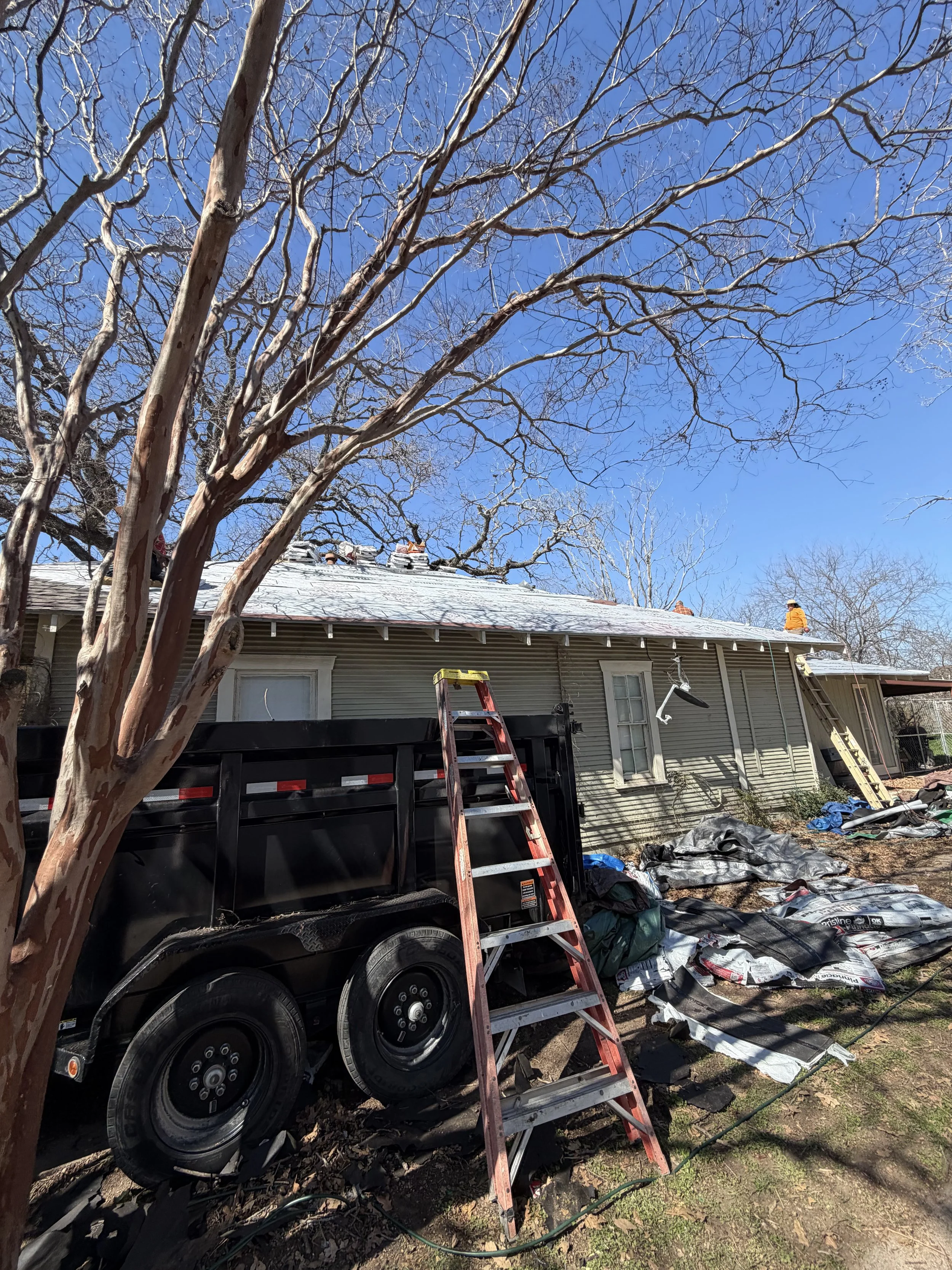 A house with a roof under construction and a worker in a yellow safety vest on the roof. Construction tools and materials are scattered around, including a red ladder leaning against the house and a black trailer in the yard. Bare trees and a clear blue sky