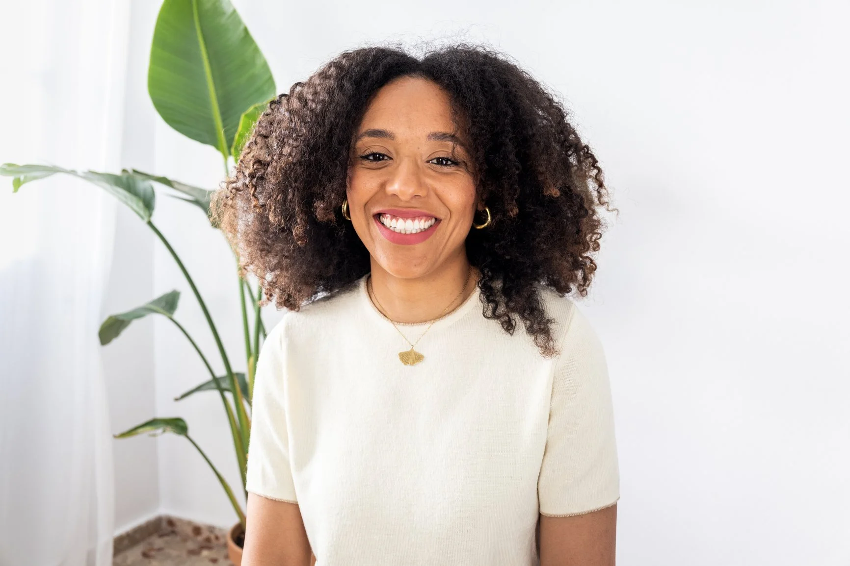 A smiling woman standing in front of a white background and a potted plant .