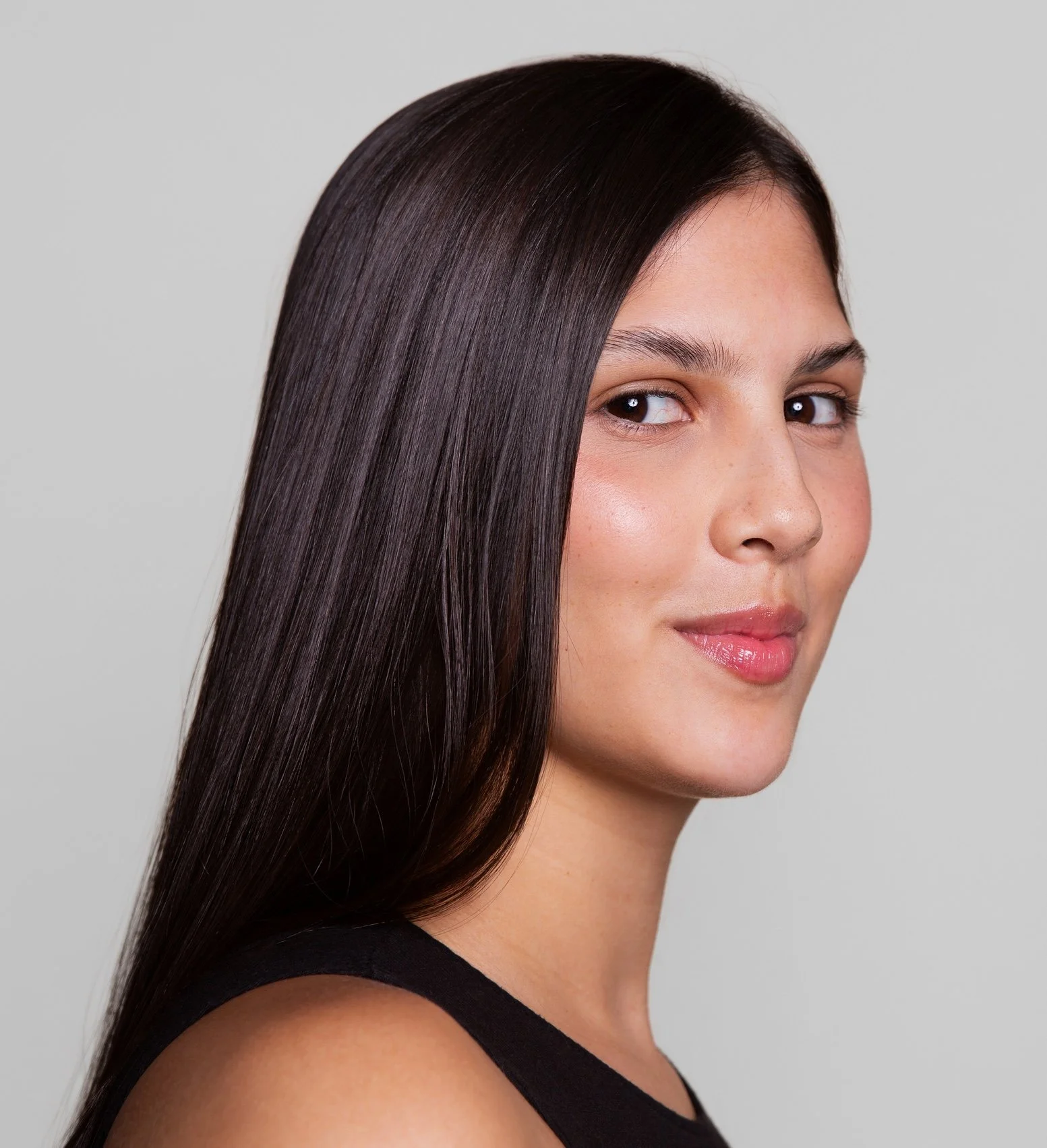 A woman with long, straight, dark brown hair, partial face, and wearing a black top, smiling slightly against a light gray background.