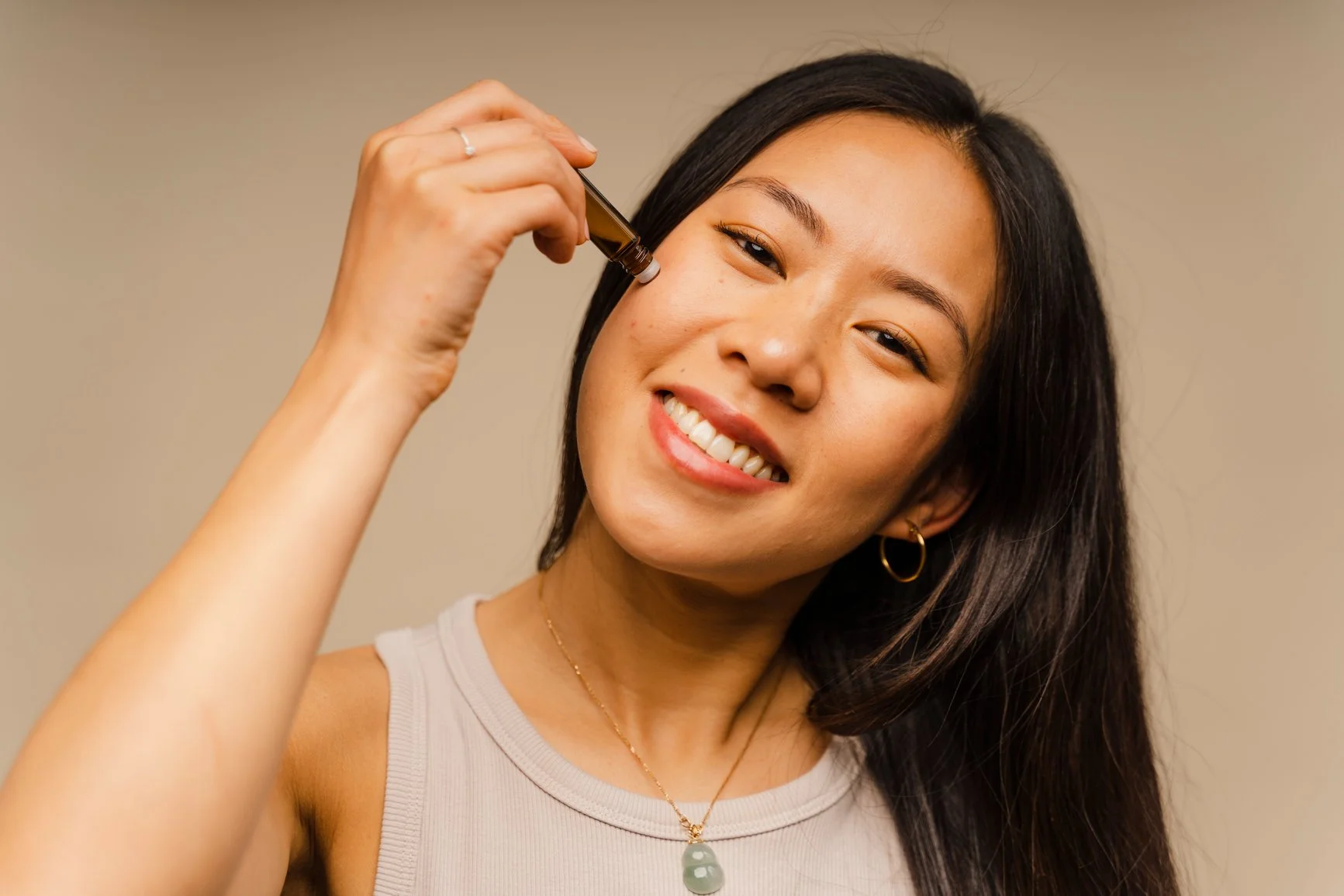 Woman with long dark hair smiling and holding a dropper with liquid near her face.