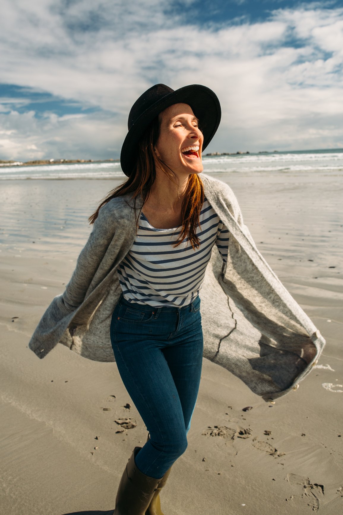 A woman walking on the beach and smiling happily with the ocean and cloudy sky in the background.