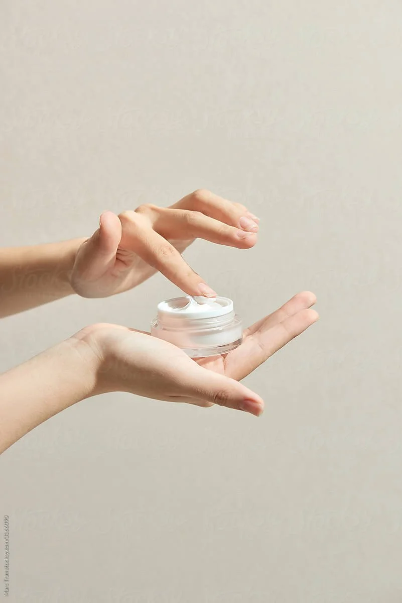 Hands holding a small jar of white cream, with one hand dipping a finger into the cream.