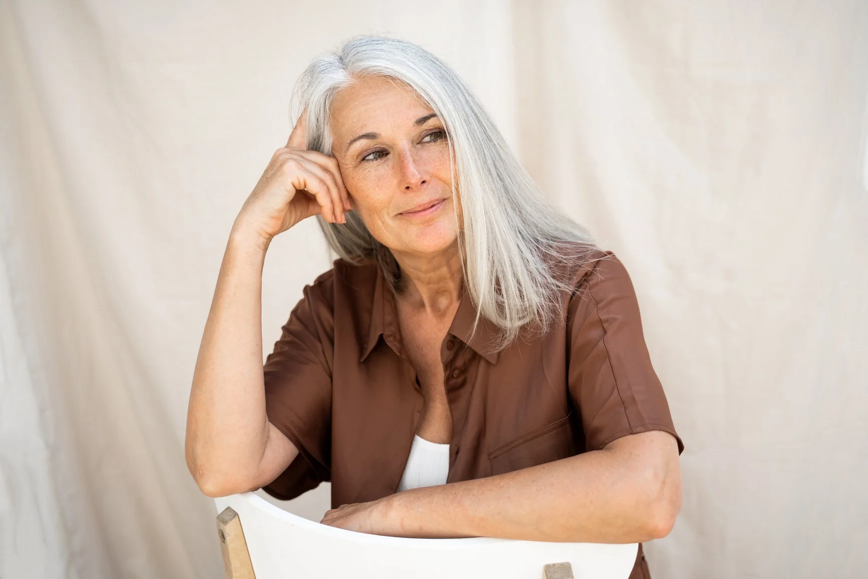 A thoughtful, mature woman with gray hair sits at a table, resting her head on her hand.