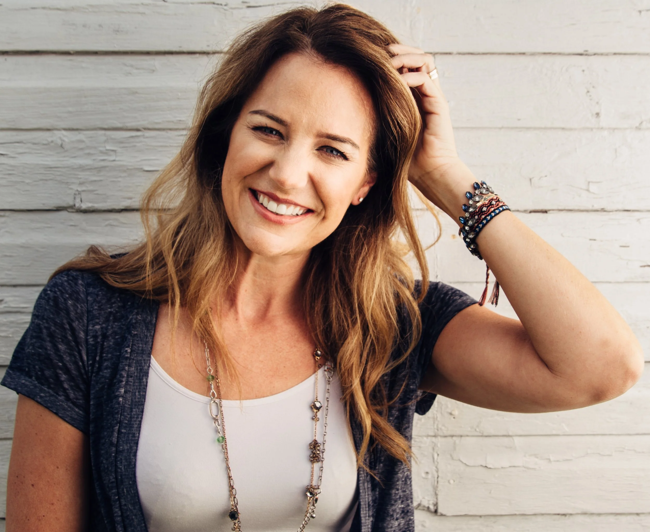 A woman with long, reddish-brown hair smiling and touching her head, standing in front of a white wooden wall. She is wearing a white top, layered necklaces, and multiple bracelets.