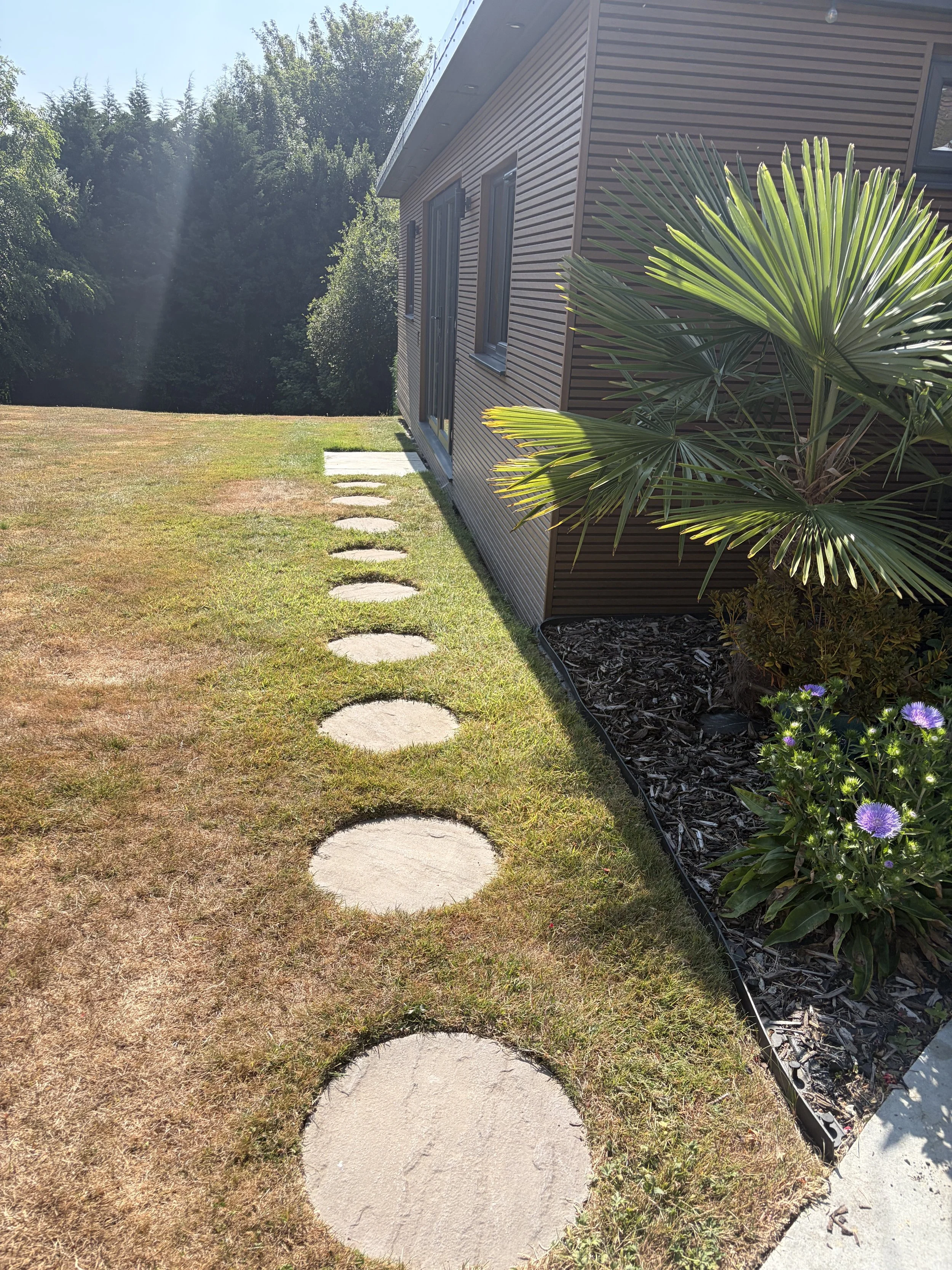 Side view of a house with brown siding, a sliding glass door, and windows. There's a small garden with a palm tree and purple flowers. A stepped pathway with circular concrete pavers cuts through the grass in the yard.