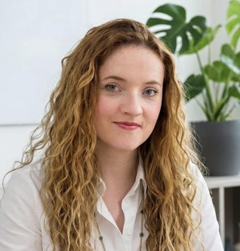 A woman with long, curly brown hair and blue eyes, wearing a white shirt, sitting indoors with a large green plant in the background.