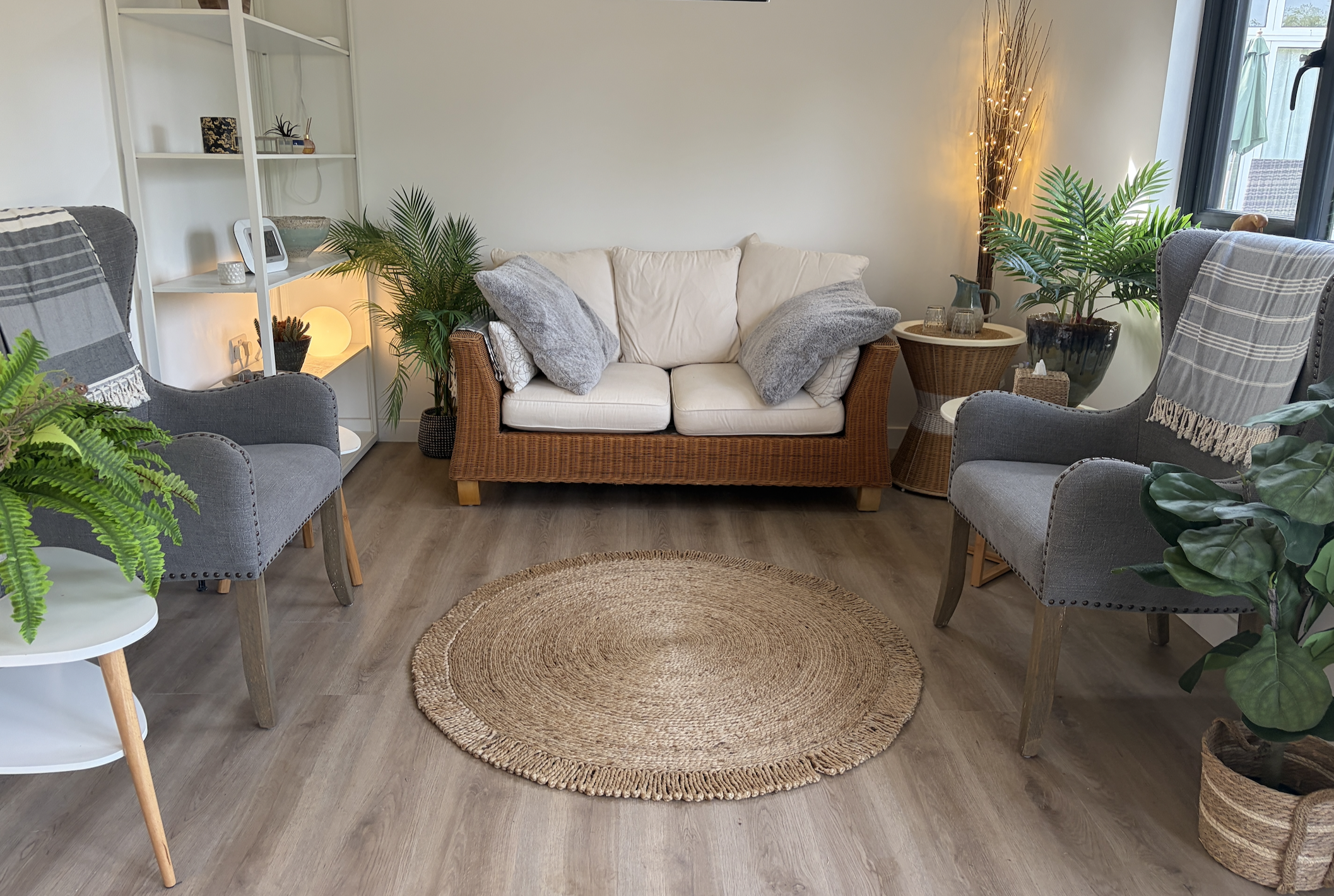 Living room with a beige sofa, grey armchairs, potted plants, a woven rug, and decorative items on shelves and tables, with natural light coming through large windows.