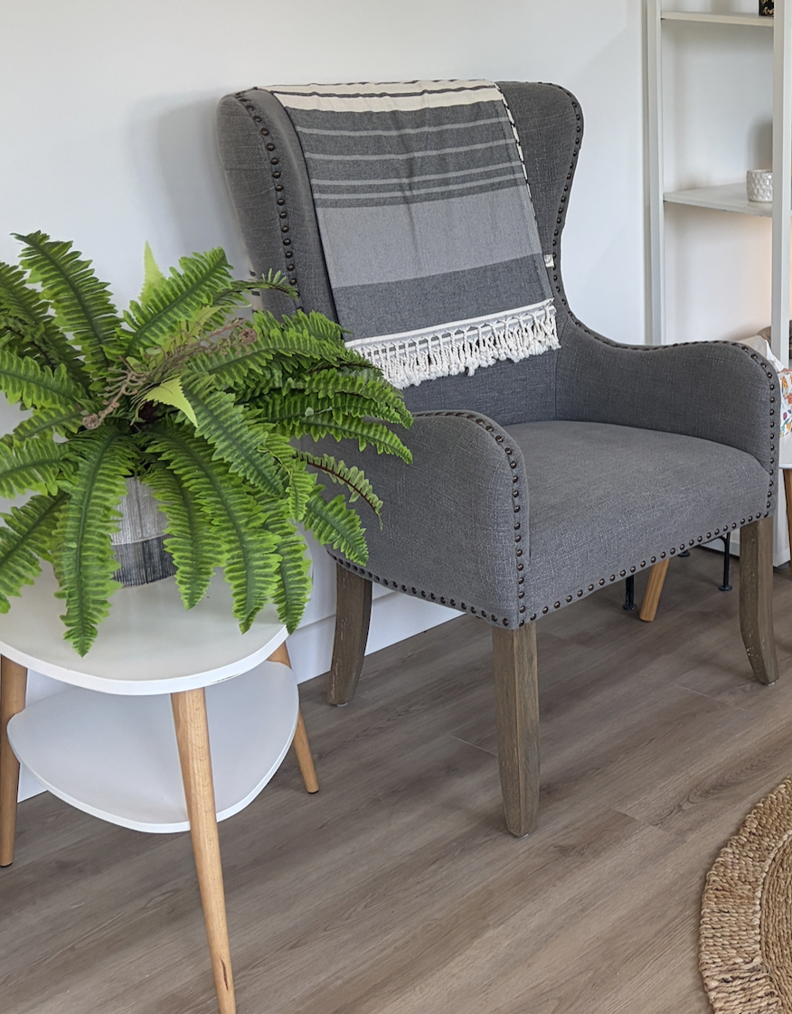 A grey upholstered armchair with wooden legs, a grey and white striped throw blanket draped over it, beside a white side table with a large potted fern plant on top, in a cozy living room.