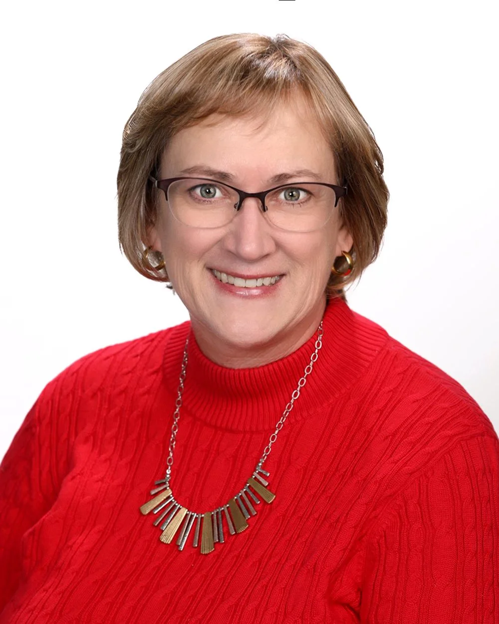 A woman with short, light brown hair and glasses, wearing a red sweater with a necklace and earrings, smiling at the camera against a white background.